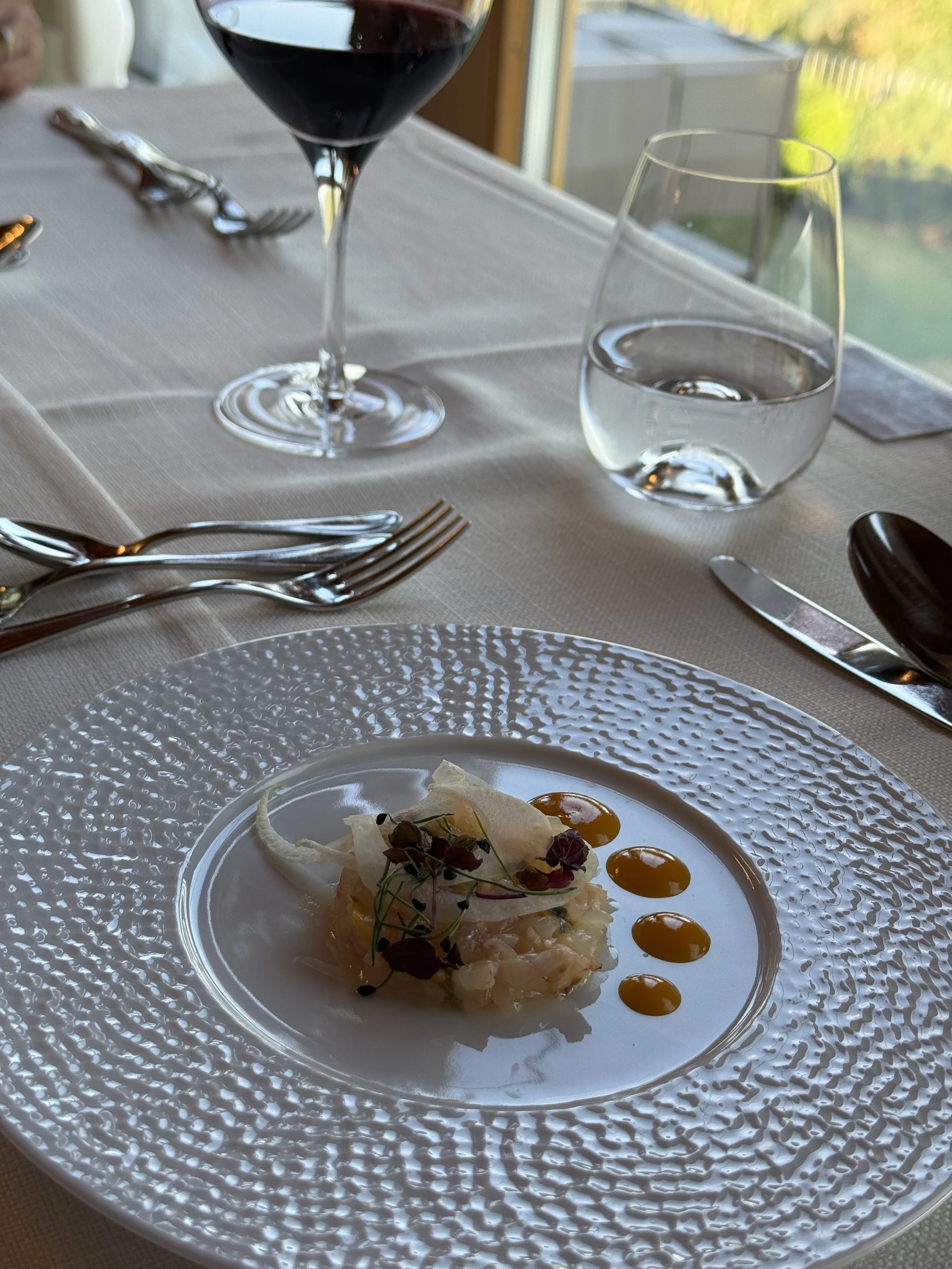 Elegant table setting with a white textured plate holding a gourmet appetizer garnished with microgreens and small edible flowers, accompanied by several drops of yellow sauce. In the background, there is a glass of red wine, a glass of water, and cu