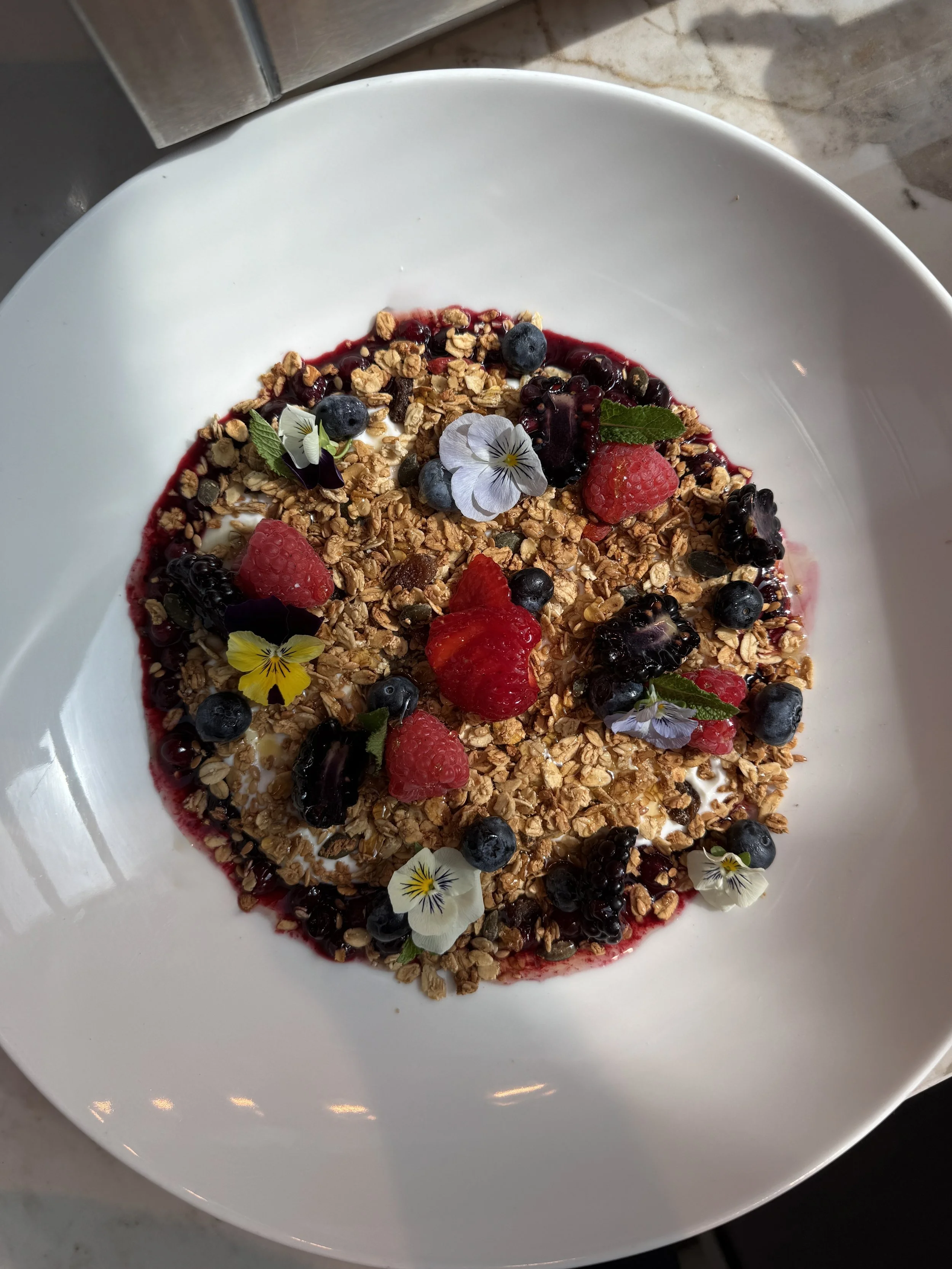 A bowl of yogurt topped with granola, mixed berries, edible flowers, and fresh mint leaves.