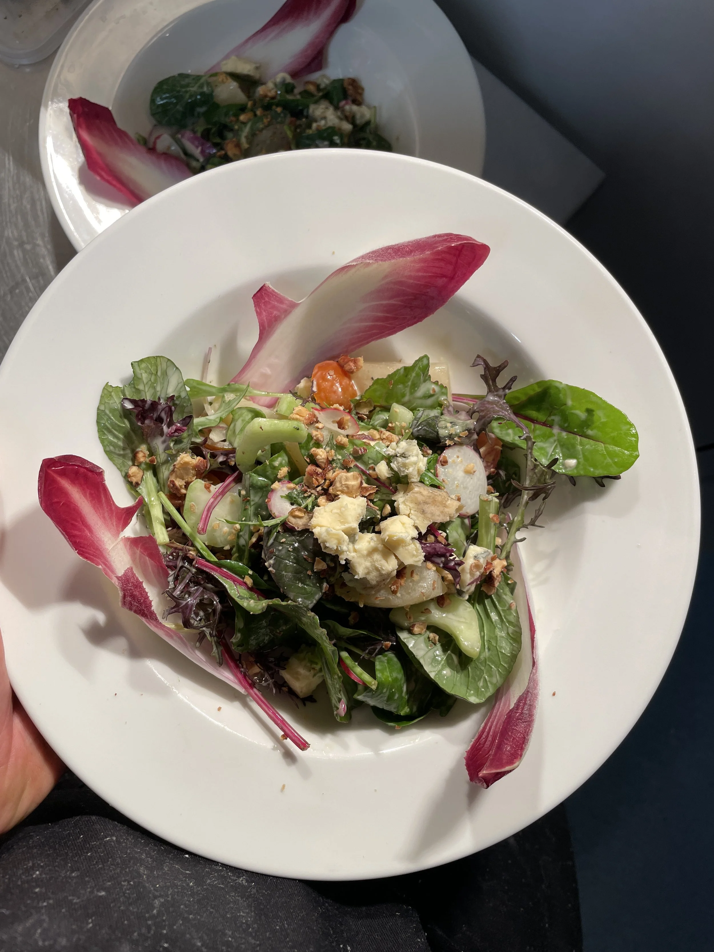 A colorful mixed salad with greens, radish slices, crumbled cheese, nuts, and radicchio leaves in a white bowl.