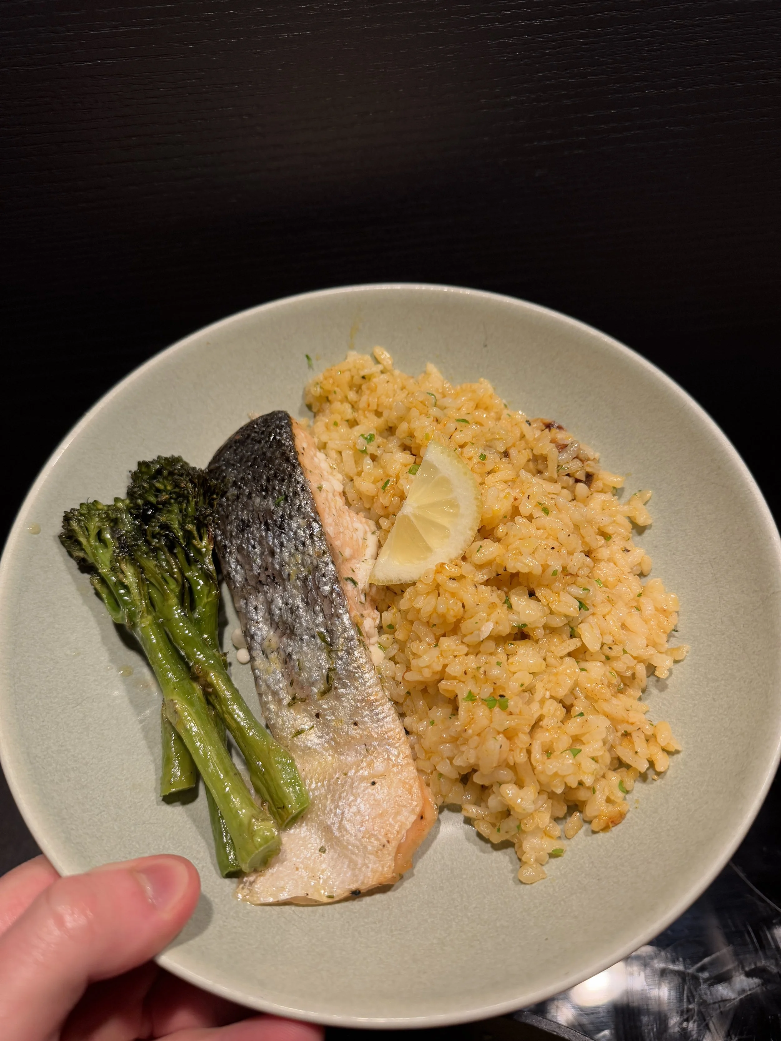 A plate of cooked rice with herbs and spices, a baked salmon fillet with crispy skin, steamed broccolini, and a lemon wedge.