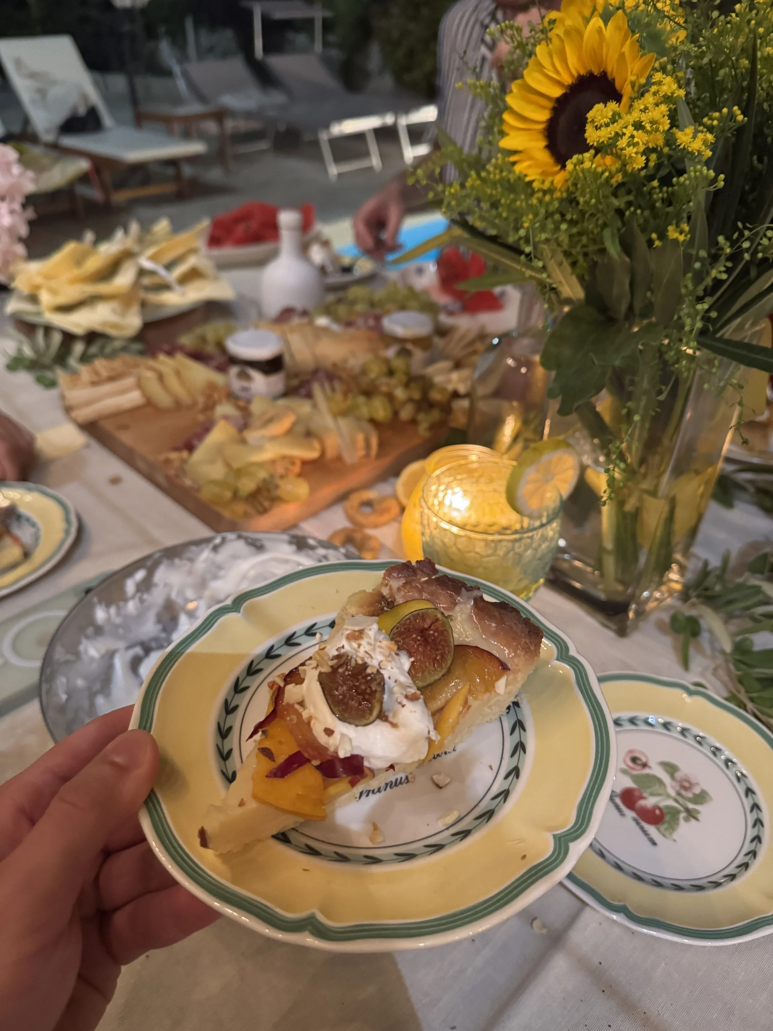 A slice of fruit tart with whipped cream, figs, and yellow fruit on a decorative plate held by a hand, with a table of food and a vase of sunflowers in the background.