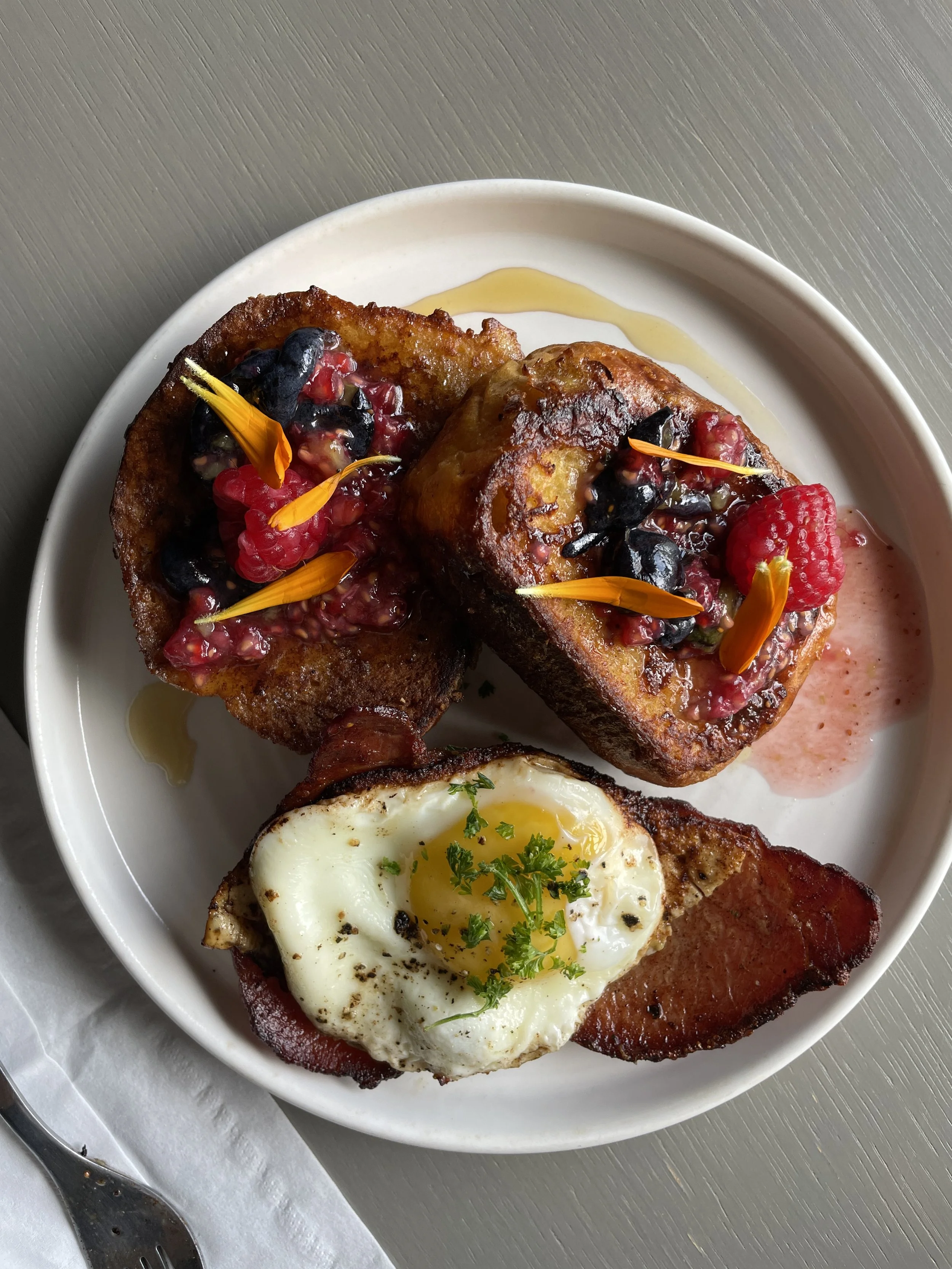 Plate of French toast topped with mixed berries, edible flower petals, and syrup, with a sunny-side-up egg garnished with herbs and crispy bacon