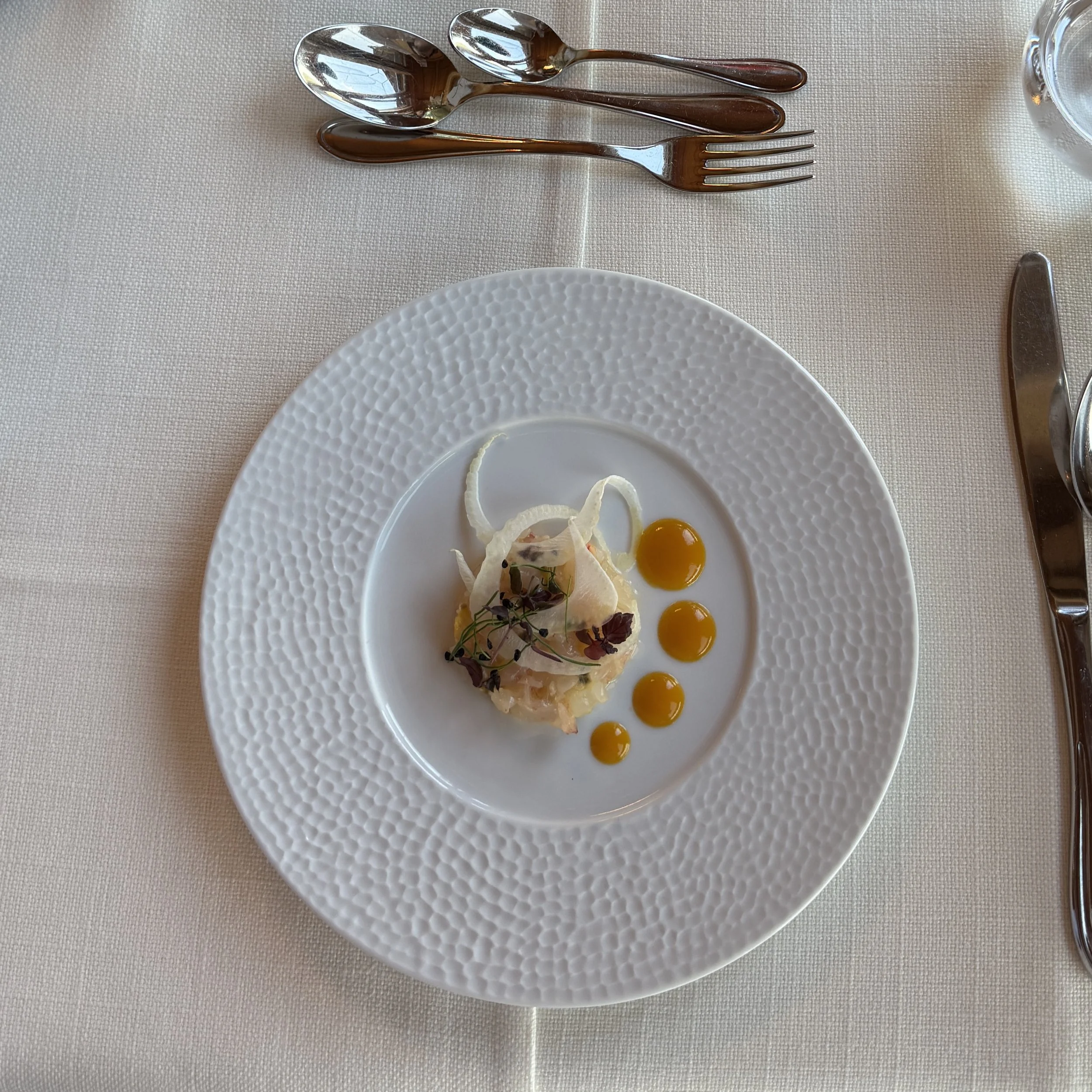 Elegant plated dish with garnishes, small droplets of sauce, and microgreens on a white textured plate, set on a table with silverware and a glass of water.