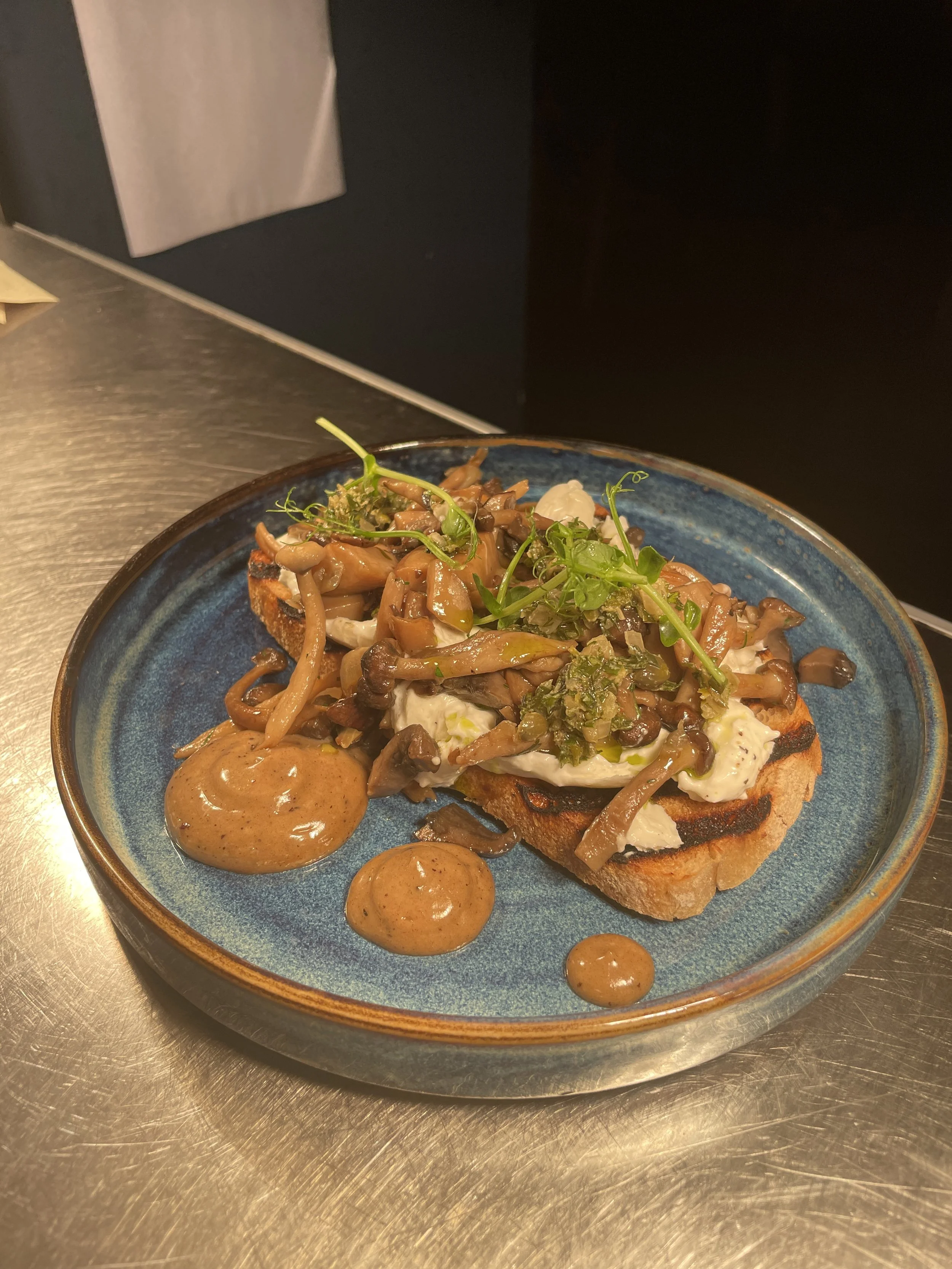 A plate with toasted bread topped with cream cheese and sautéed mushrooms, garnished with green herbs, accompanied by dollops of brown sauce on a blue ceramic plate.