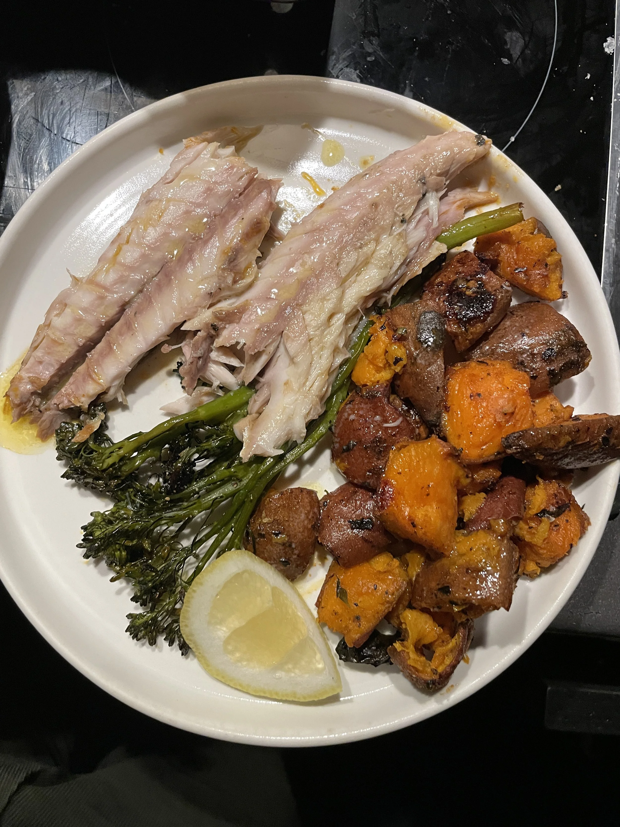Plate with cooked fish, roasted potatoes with herbs, grilled broccolini, and a lemon wedge.