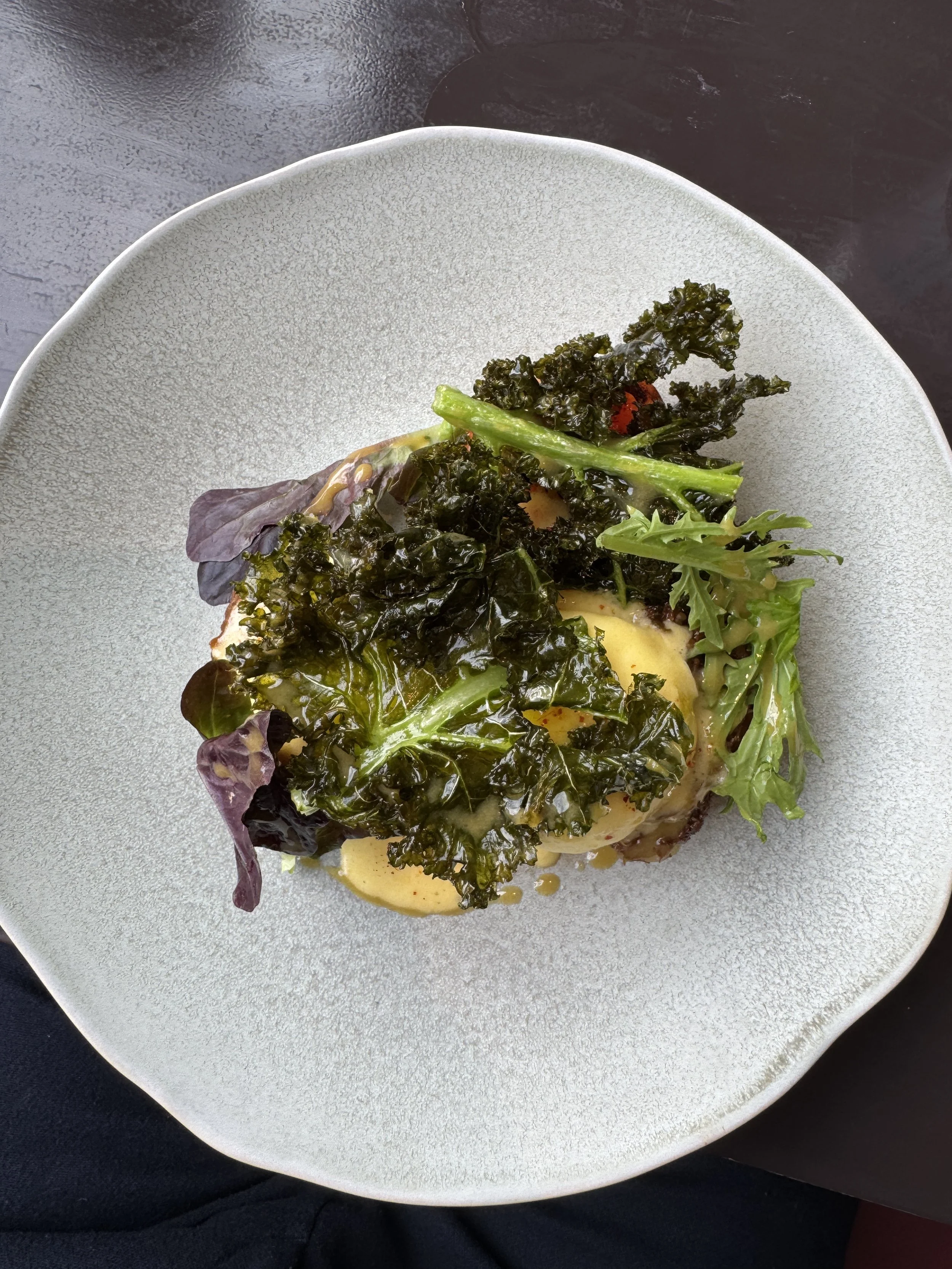 A dish with leafy greens, including kale and other mixed greens, on top of a piece of bread or toast with a creamy yellow sauce drizzle, served on a gray plate.