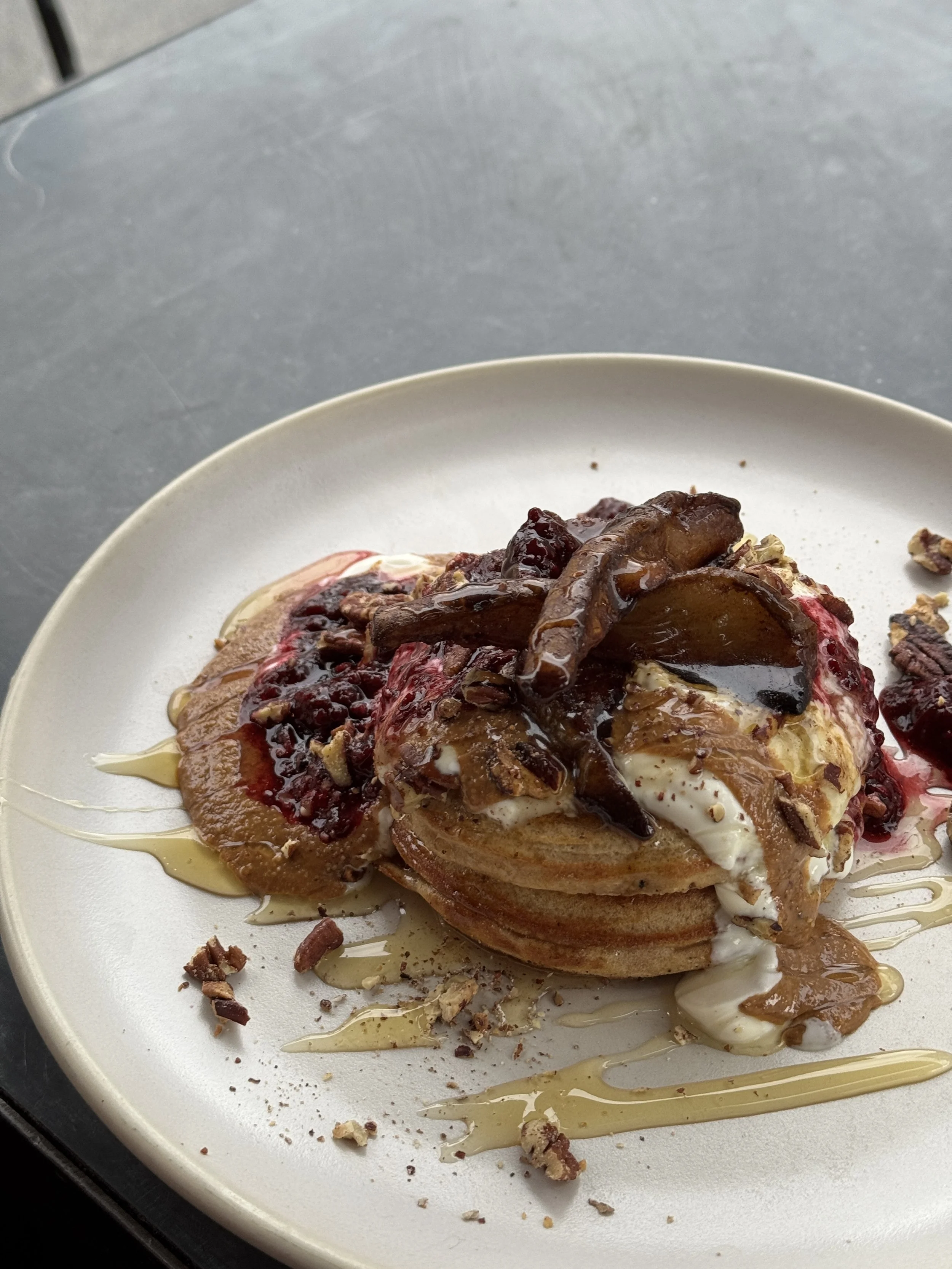 A plate with a stack of what appears to be pancakes topped with whipped cream, caramel sauce, berry compote, chopped nuts, and caramel drizzle.