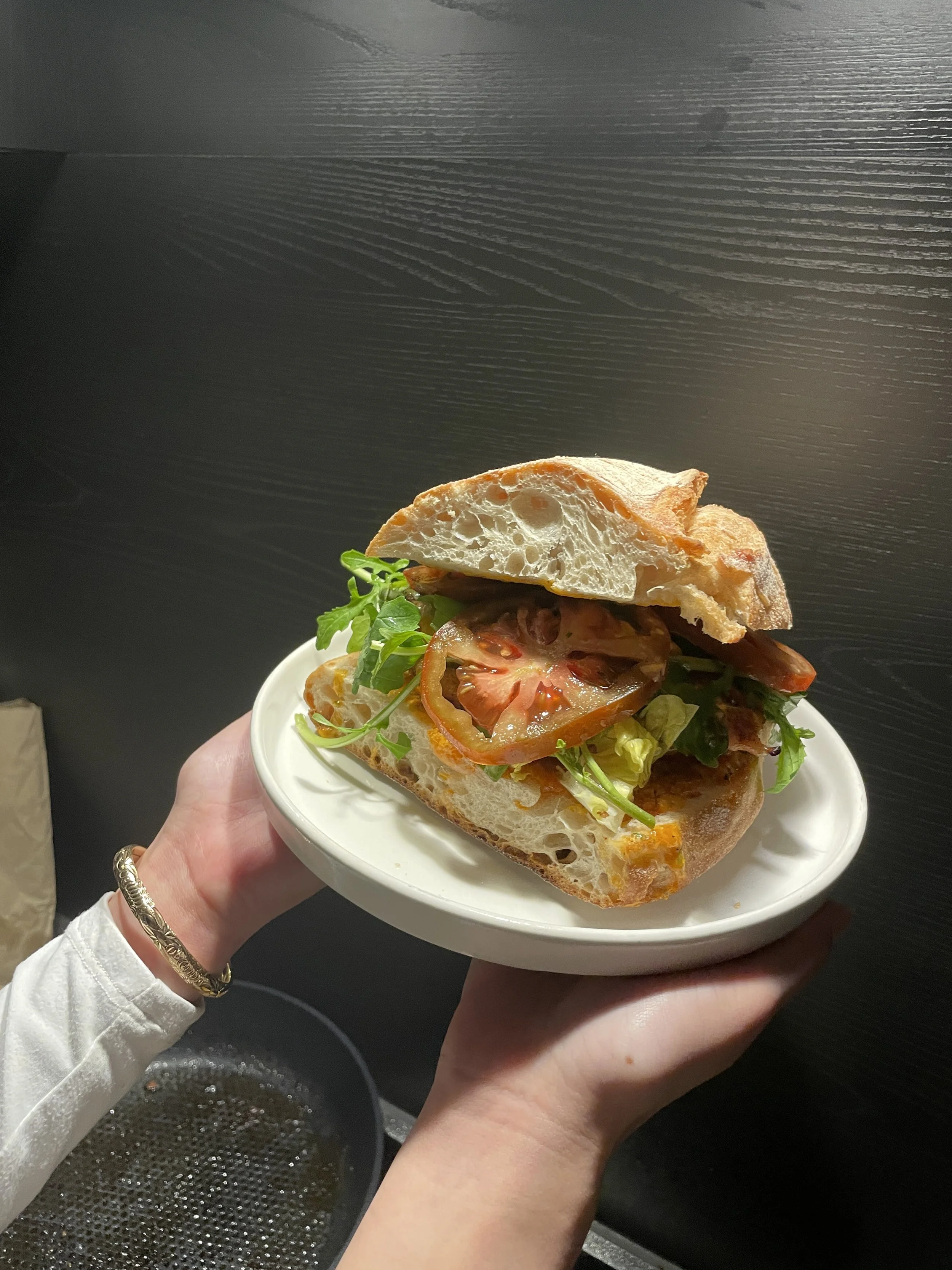 A person holding a white plate with a sandwich containing slices of tomato, lettuce, meat, and a baguette-style bread against a dark wooden background.