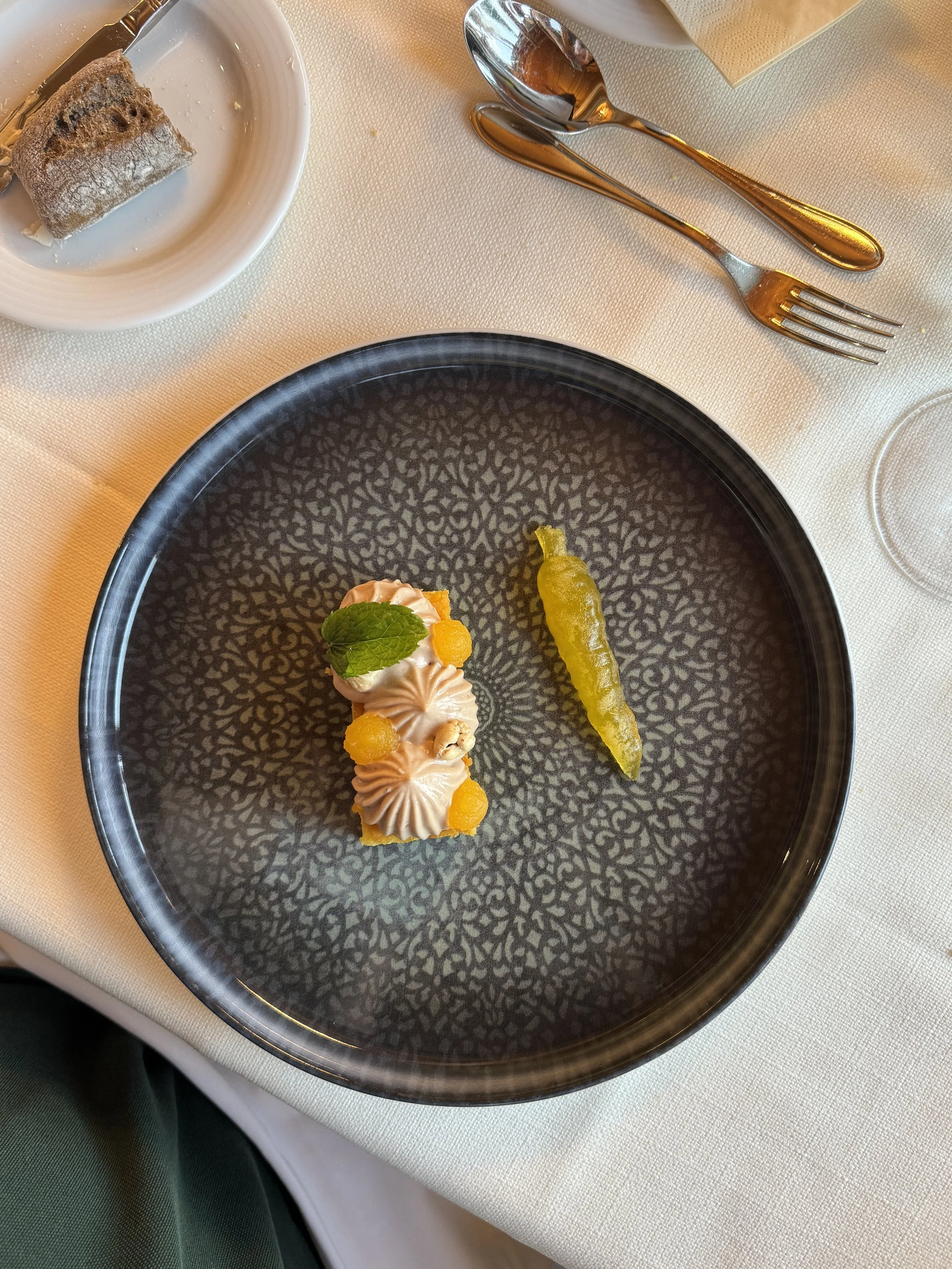 Empty black plate with a small dessert topped with cream, orange spheres, a mint leaf, and a small green pepper on the right.