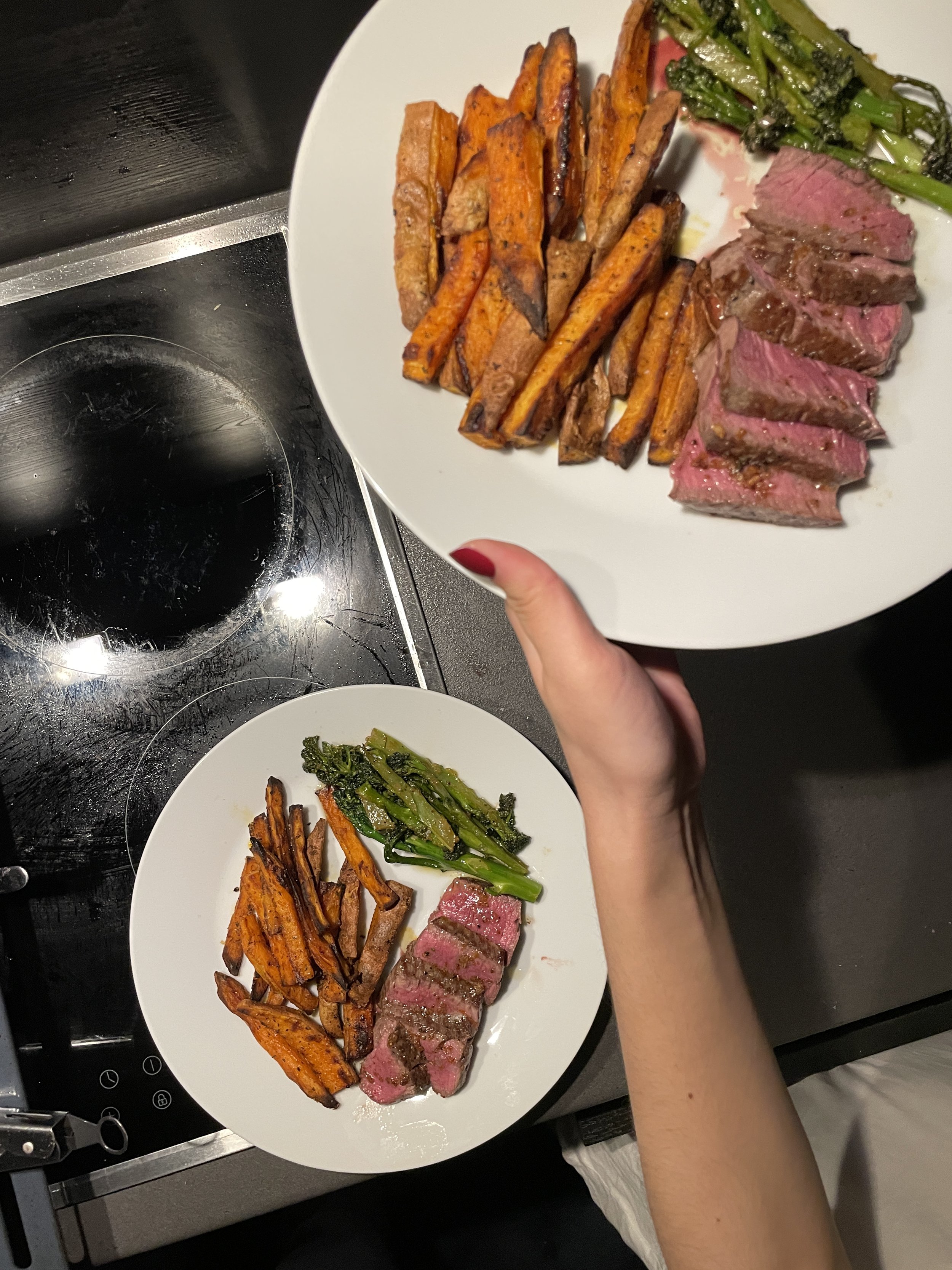 Two plates of food on a black stovetop. Each plate has grilled steak slices, roasted sweet potato fries, and sautéed green leafy vegetables.