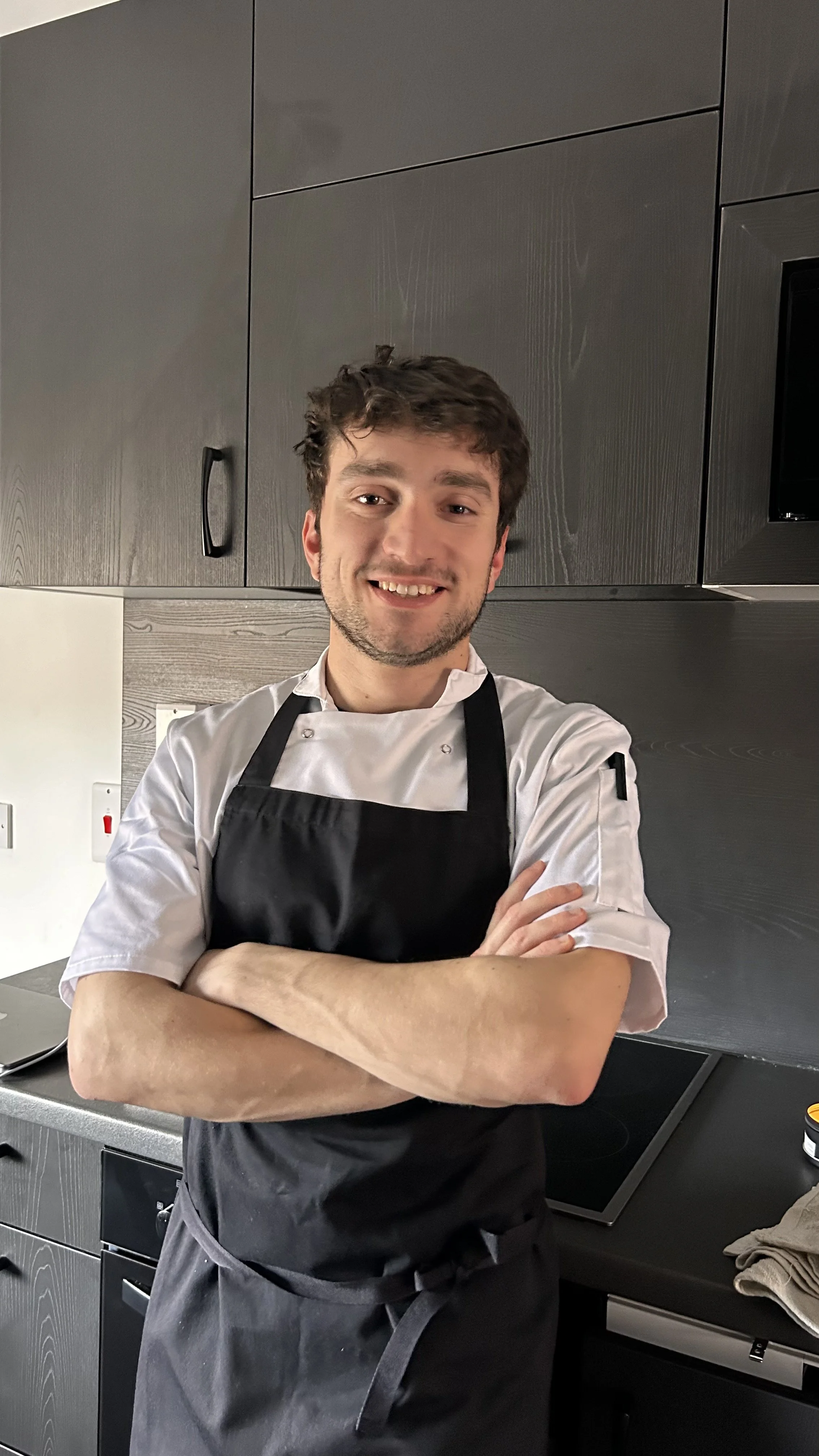 Young male chef smiling, standing crosses arms in a modern kitchen with dark cabinetry, wearing a white chef's coat and black apron.