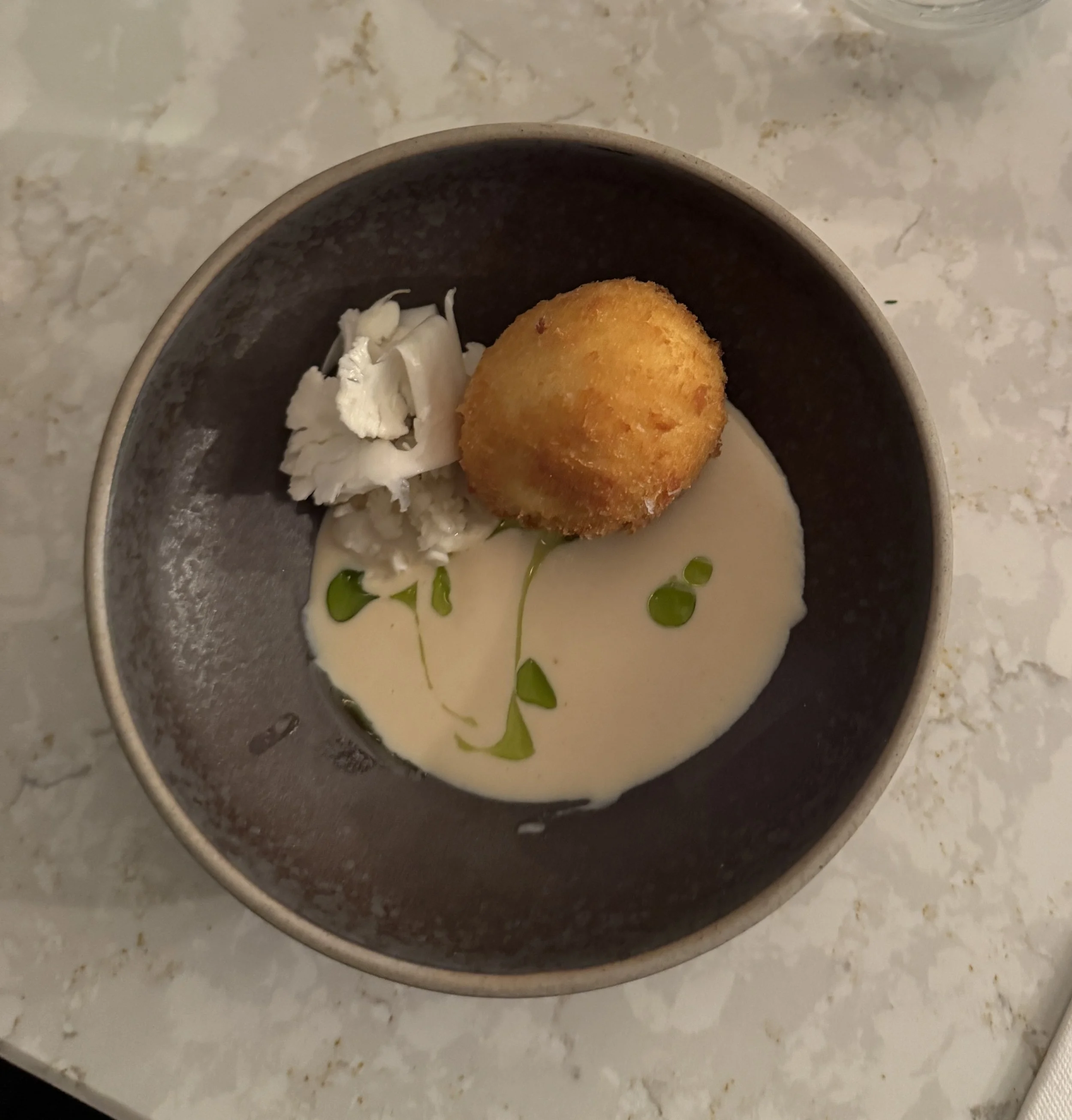 A bowl with a breaded fried ball, pieces of white fish, and a creamy sauce garnished with green oil droplets, on a marble countertop.