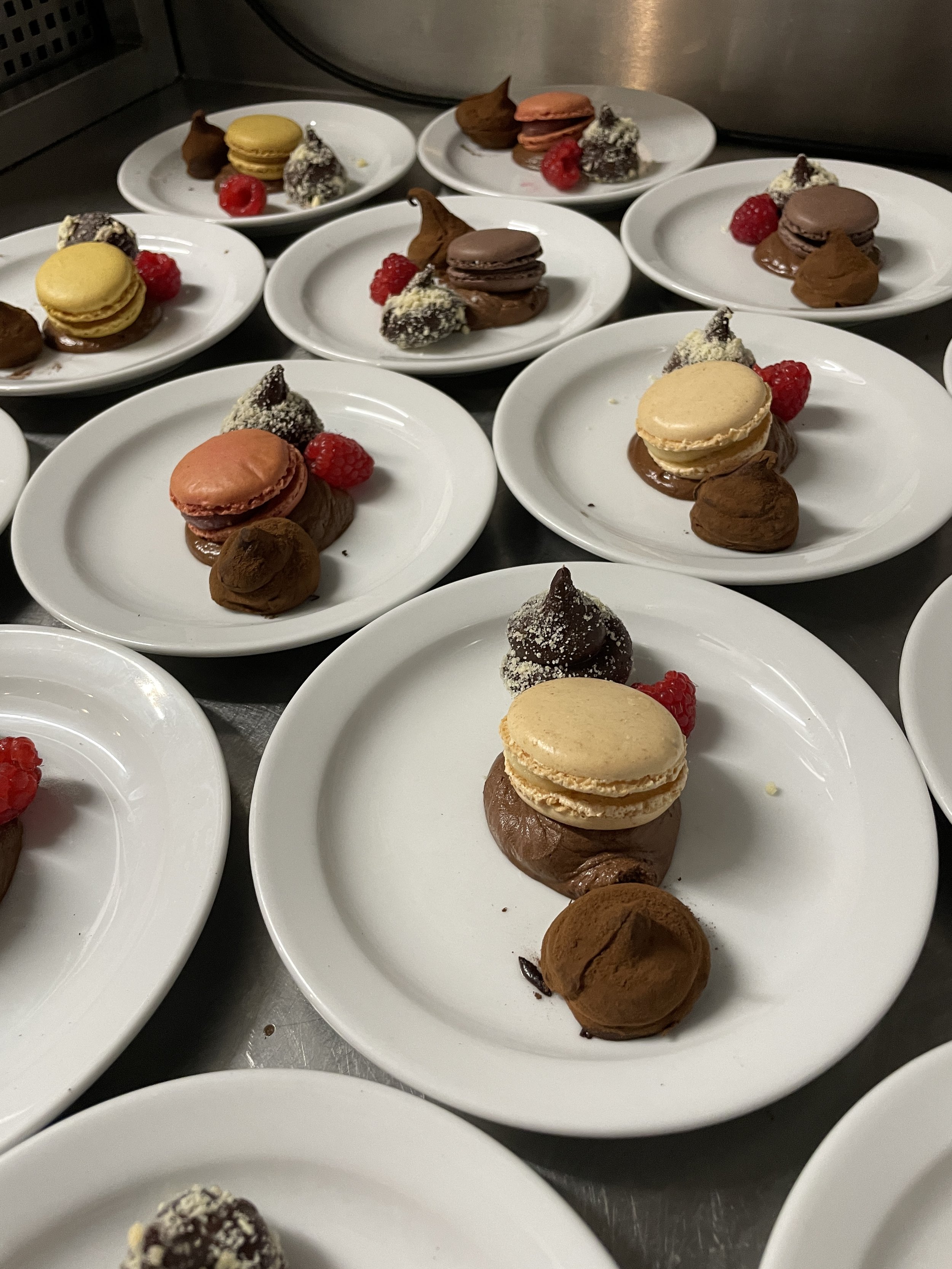 Multiple white plates with assorted desserts including macarons, chocolates, raspberry berries, and dollops of chocolate mousse or whipped cream, arranged on a stainless steel surface.
