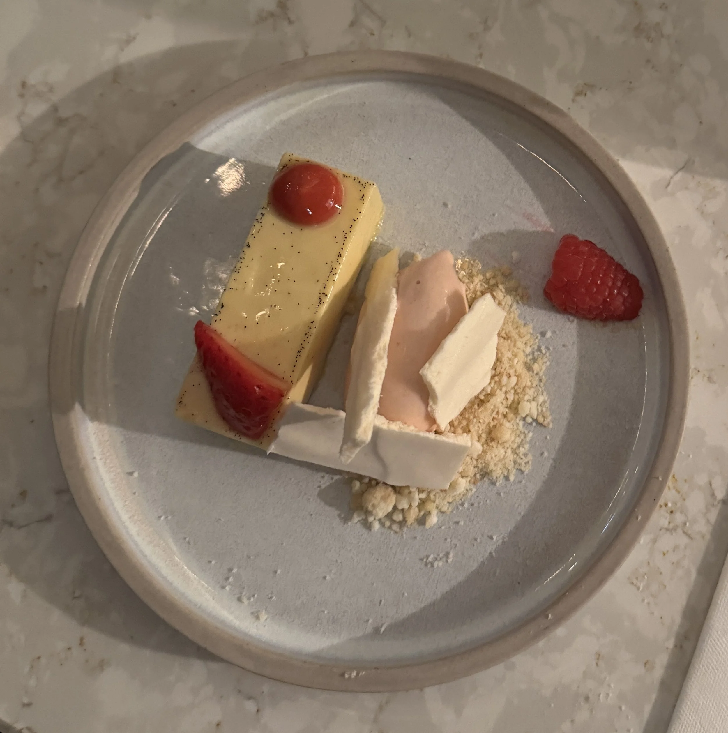 A dessert plate with assorted desserts, including a white chocolate and raspberry mousse, a piece of cheesecake with a cherry on top, a raspberry, and some powdered crumbs, all arranged on a white plate.