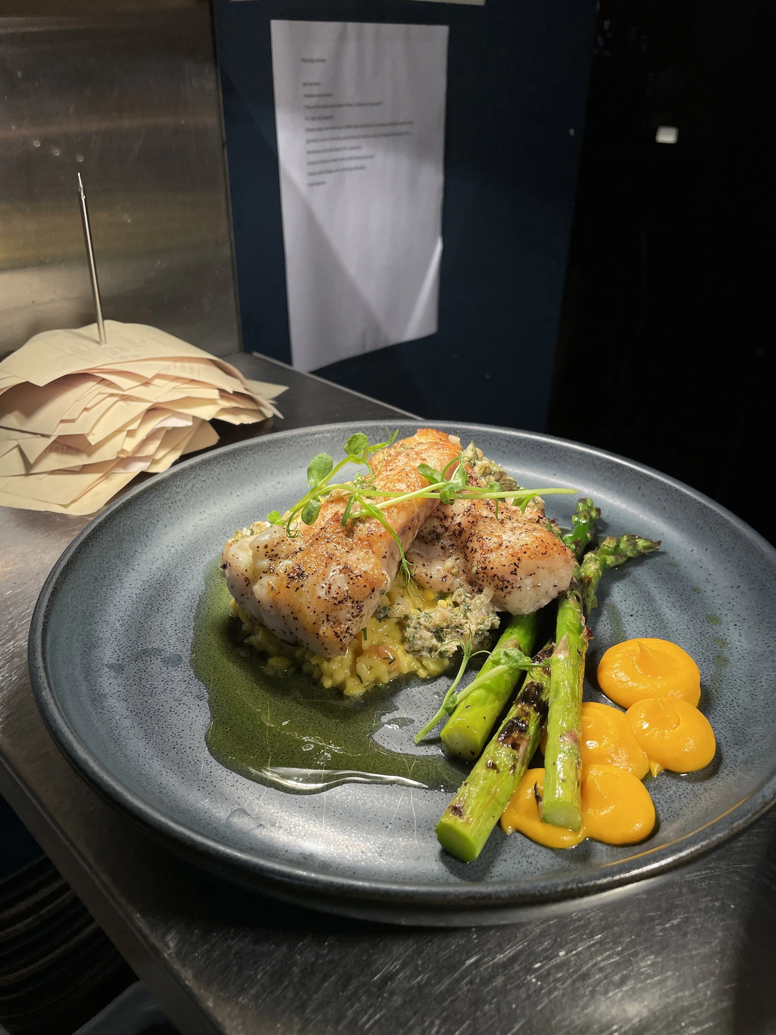 A plated dish featuring two pieces of seared fish fillets garnished with microgreens, served with grilled asparagus, creamy yellow sauce, and a bed of rice or grains.