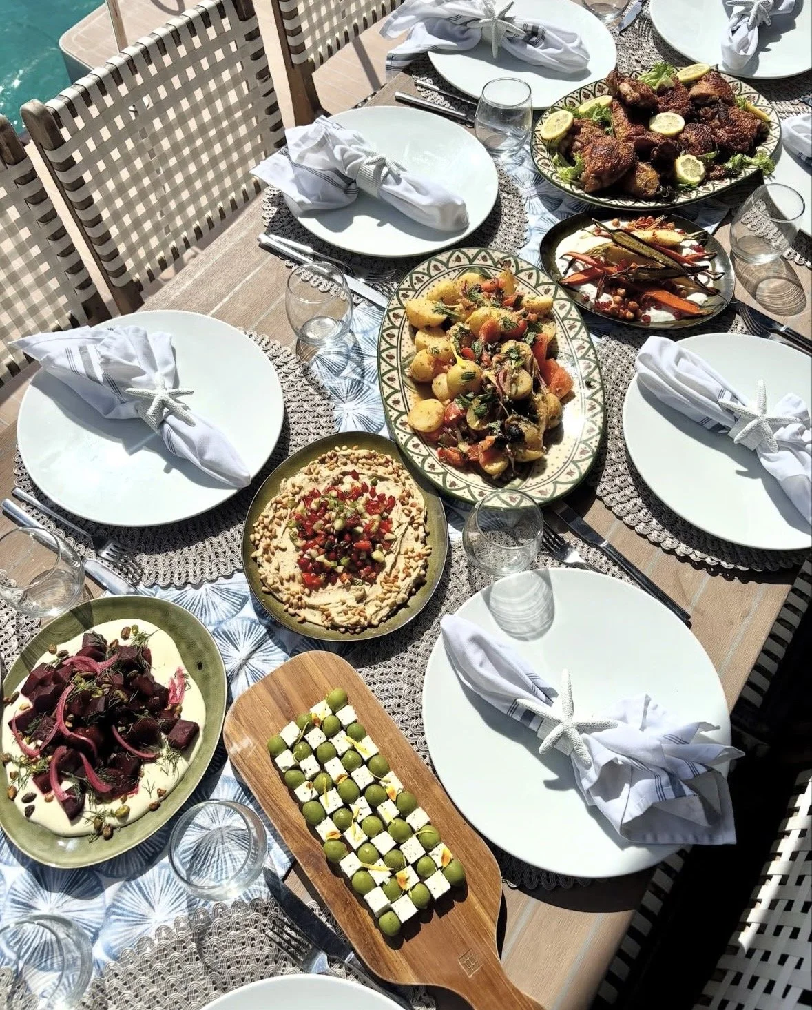 Outdoor dining table set with various dishes including salads, roasted chicken, and appetizers, with empty white plates and glasses, surrounded by chairs on a sunny day.