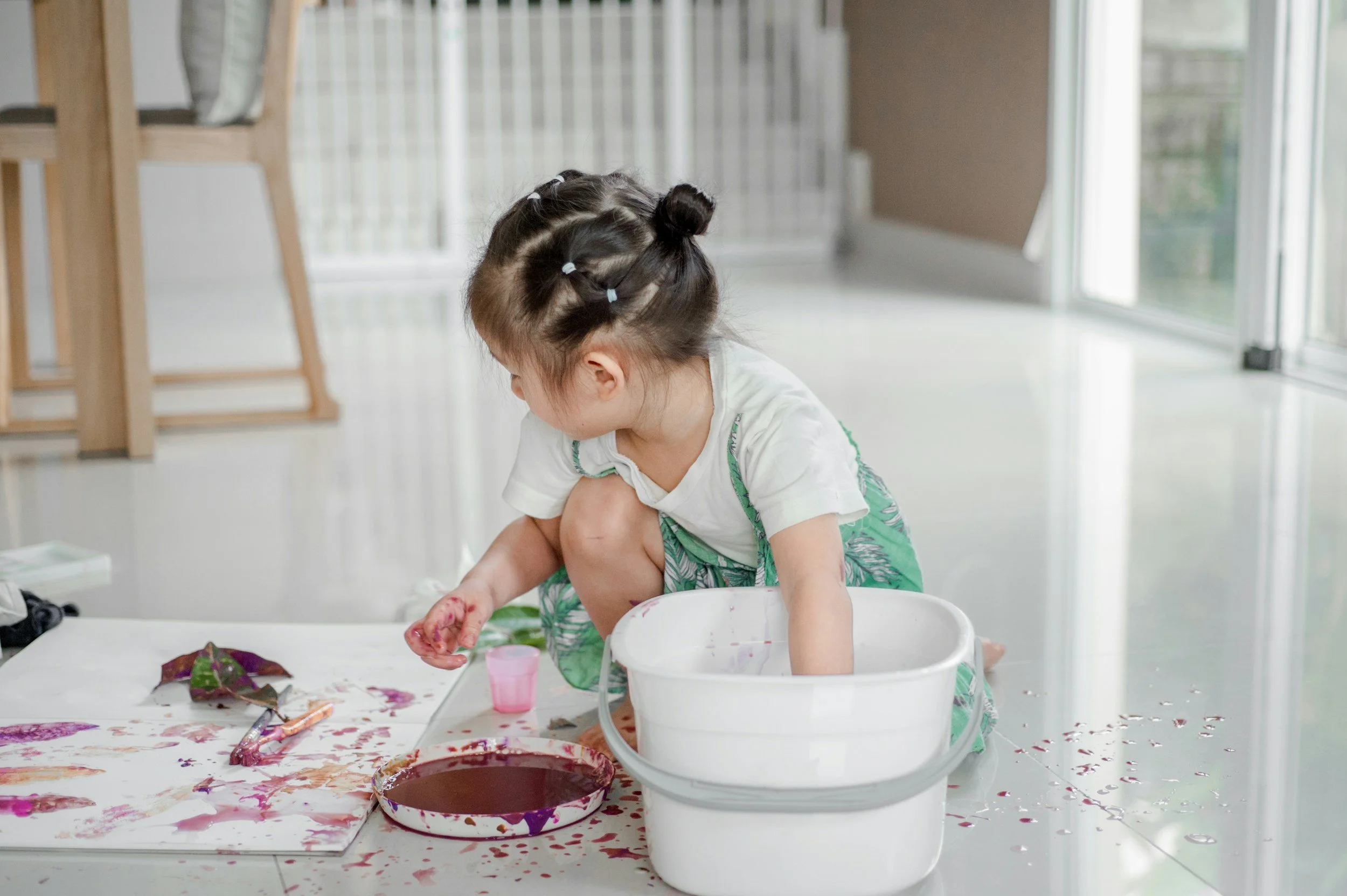 Child kneeling on the floor, finger-painting with purple and red colors on paper, surrounded by paint splatters and art supplies in a bright room.