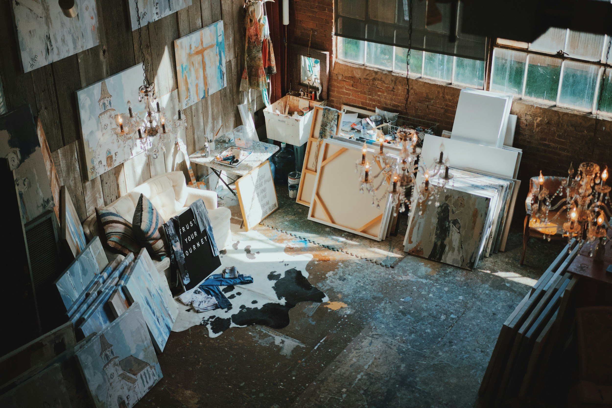 An art studio with various paintings and canvases, some leaning against a brick wall and others standing upright. The room features a cowhide rug, a cream-colored tufted sofa with pillows, chandeliers hanging from the ceiling, and a window letting in natural light. Art supplies and scattered objects are visible on tables and the floor.