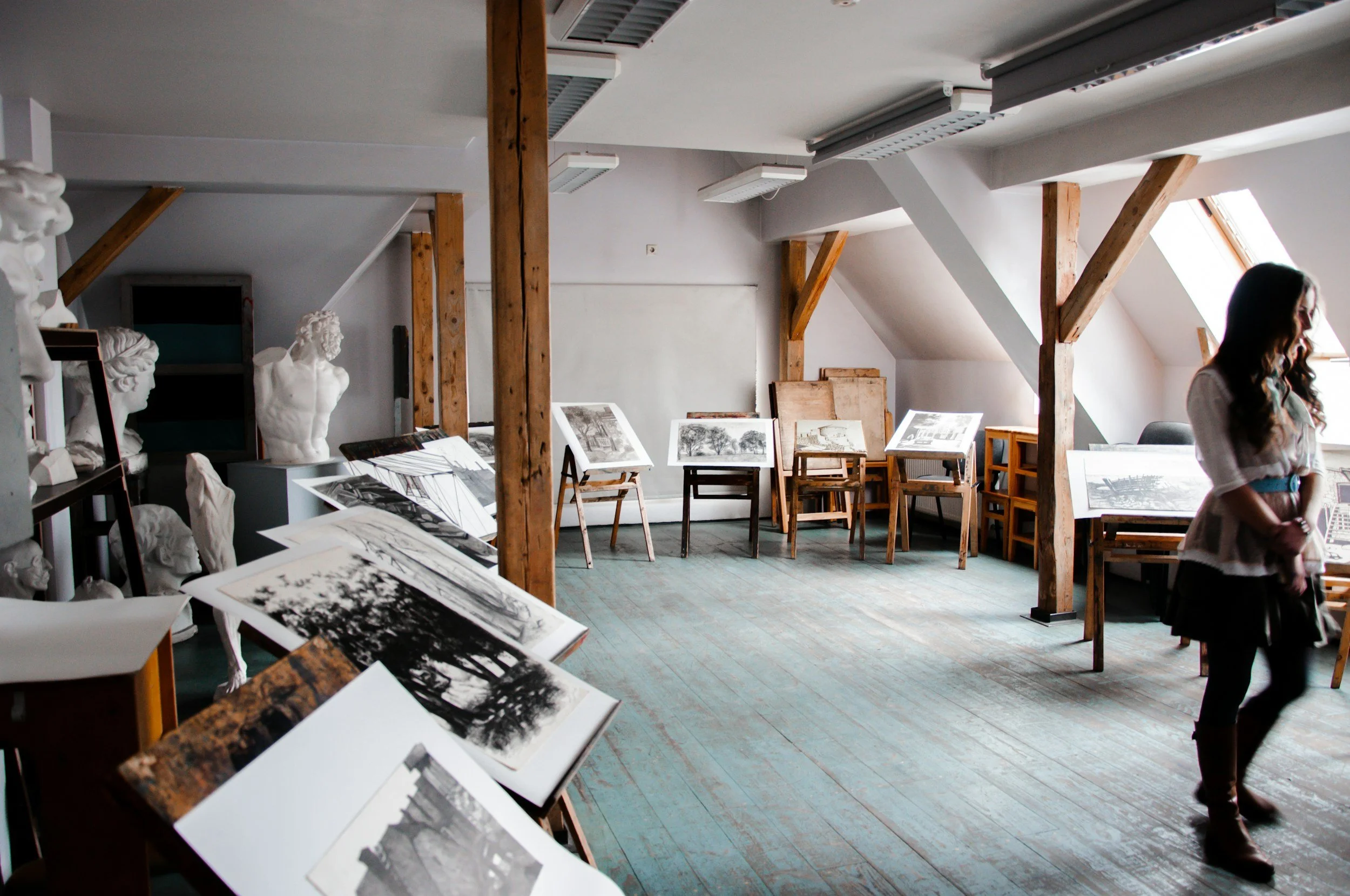 Gallery room with wooden beams, chairs, and tables displaying black and white photographs and sketches, with sculptures on the left and a woman walking on the right.