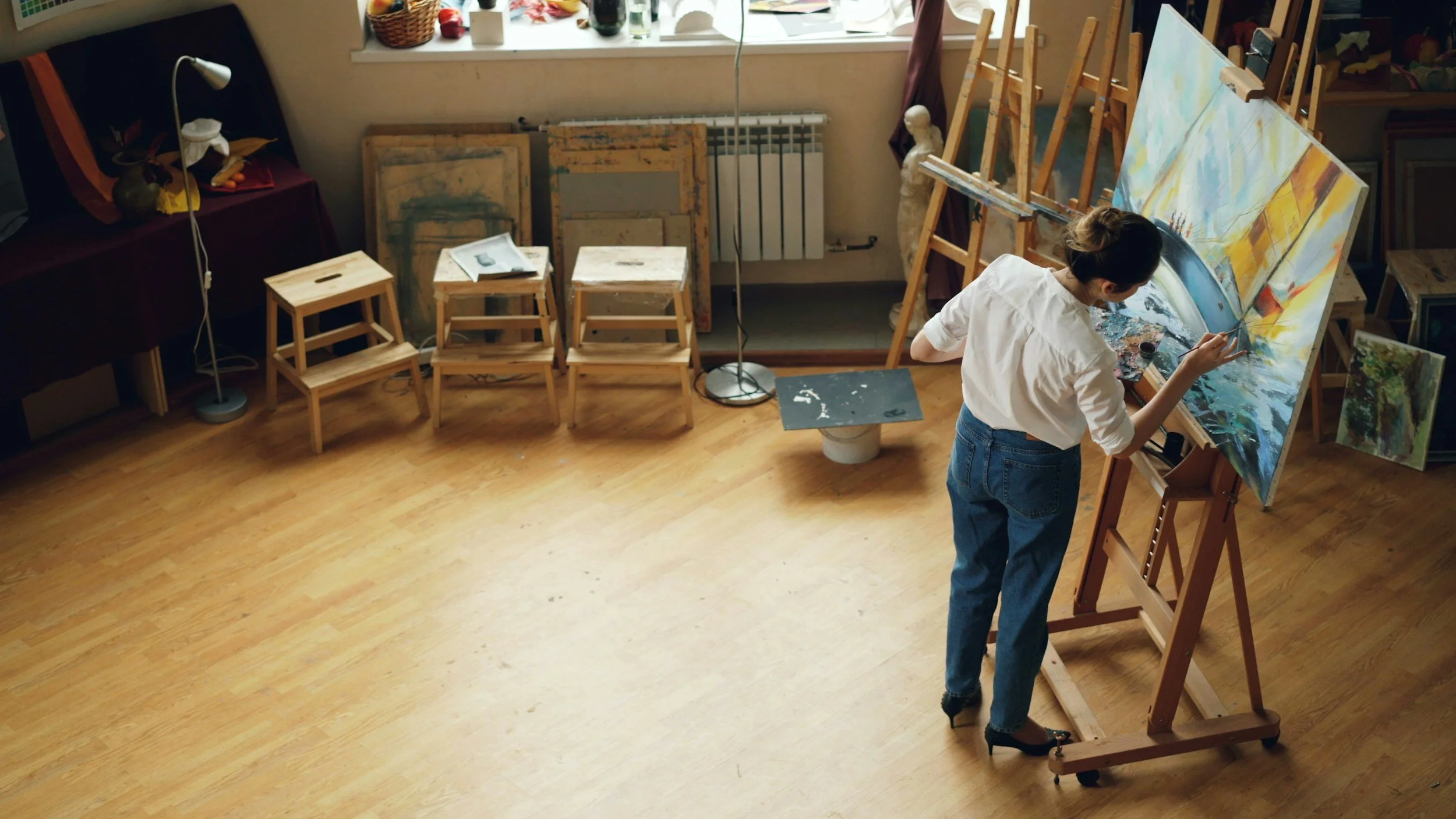 An artist painting on a large canvas in an art studio with wooden floors, multiple empty chairs, easels, and art supplies.