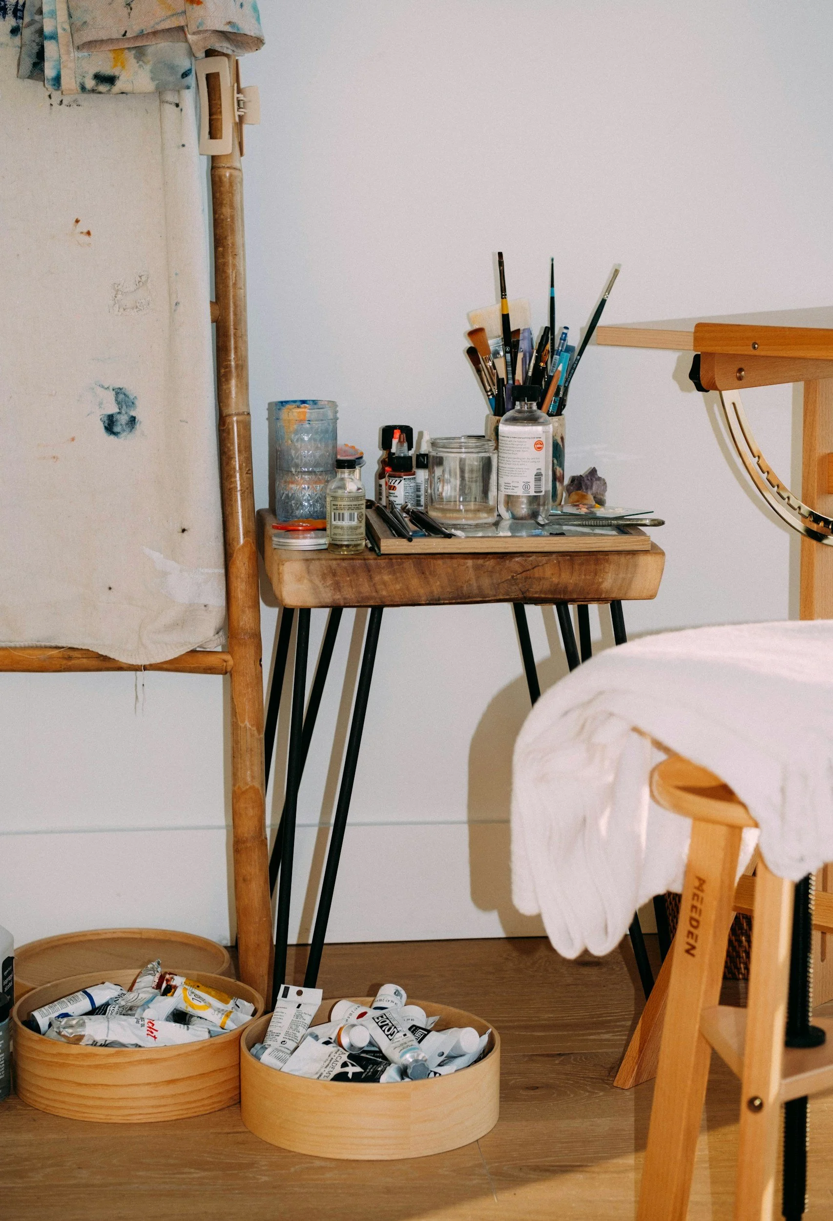 An artist's workspace with a wooden table holding paintbrushes, jars, and art supplies, and two round wooden containers filled with paint tubes on the floor.