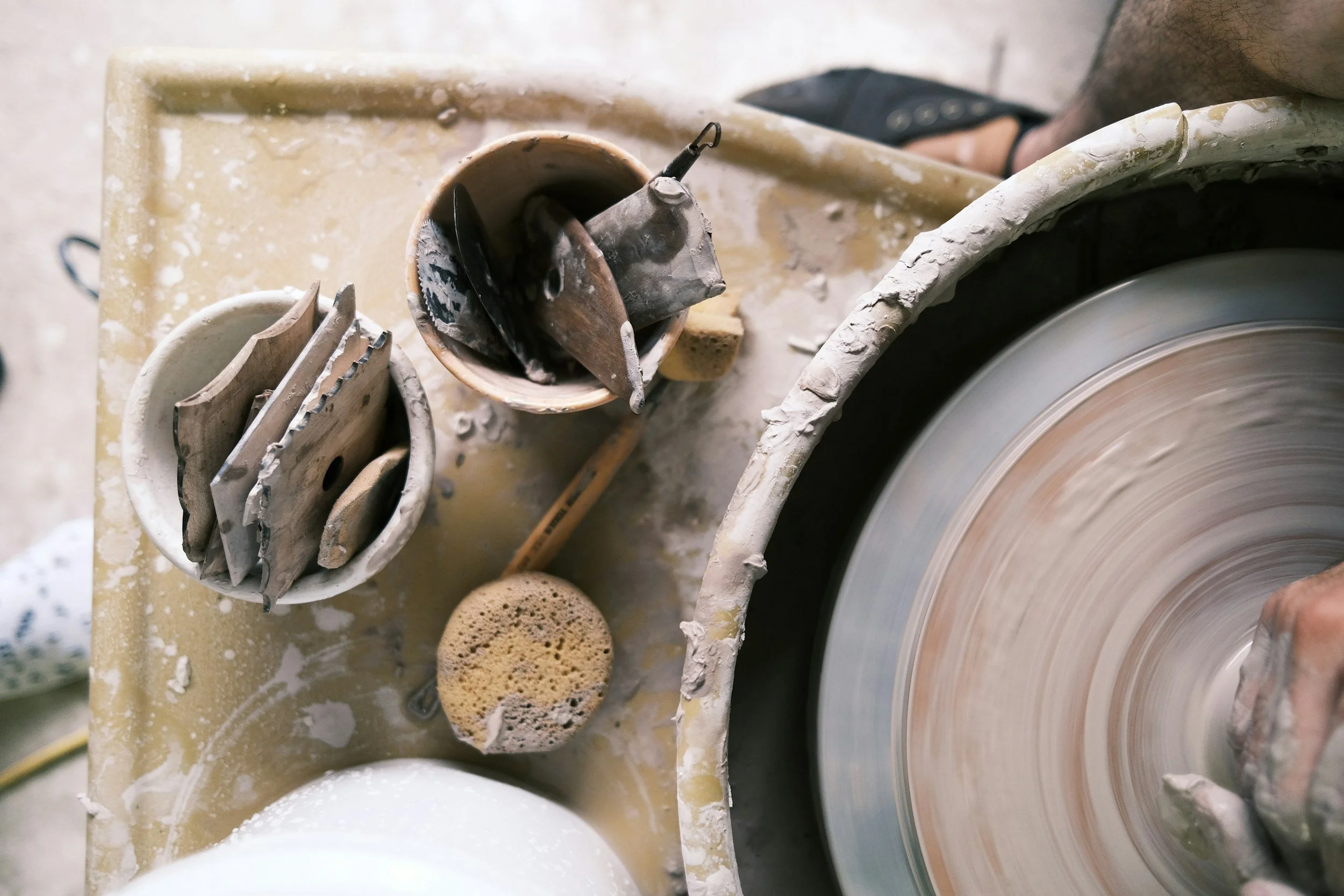 Close-up of a pottery wheel spinning clay and various pottery tools, sponge, and sponges on a messy table.