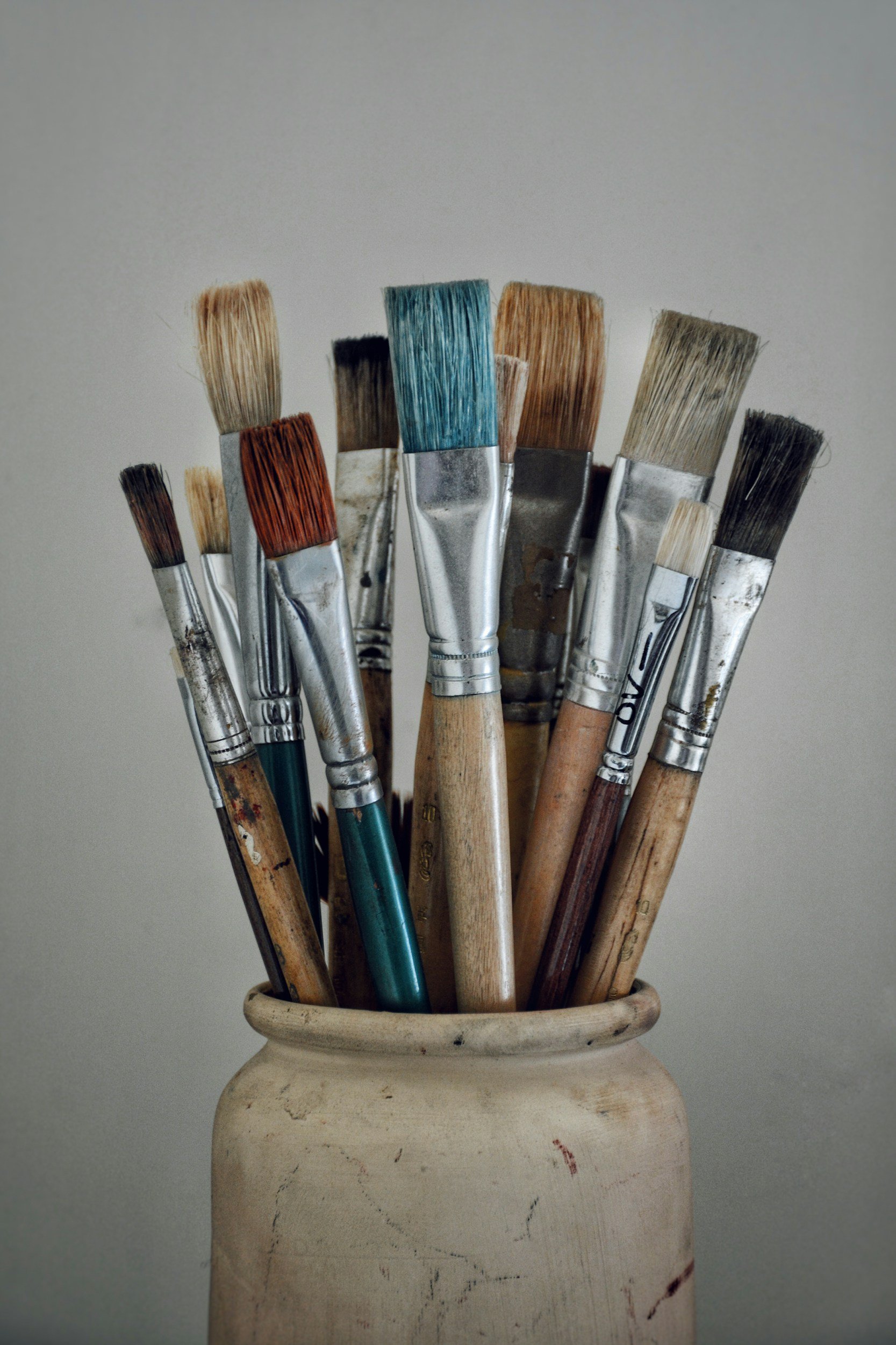 A collection of used paintbrushes with wooden and metal handles, some with paint stains, in a rustic ceramic vase against a plain background.