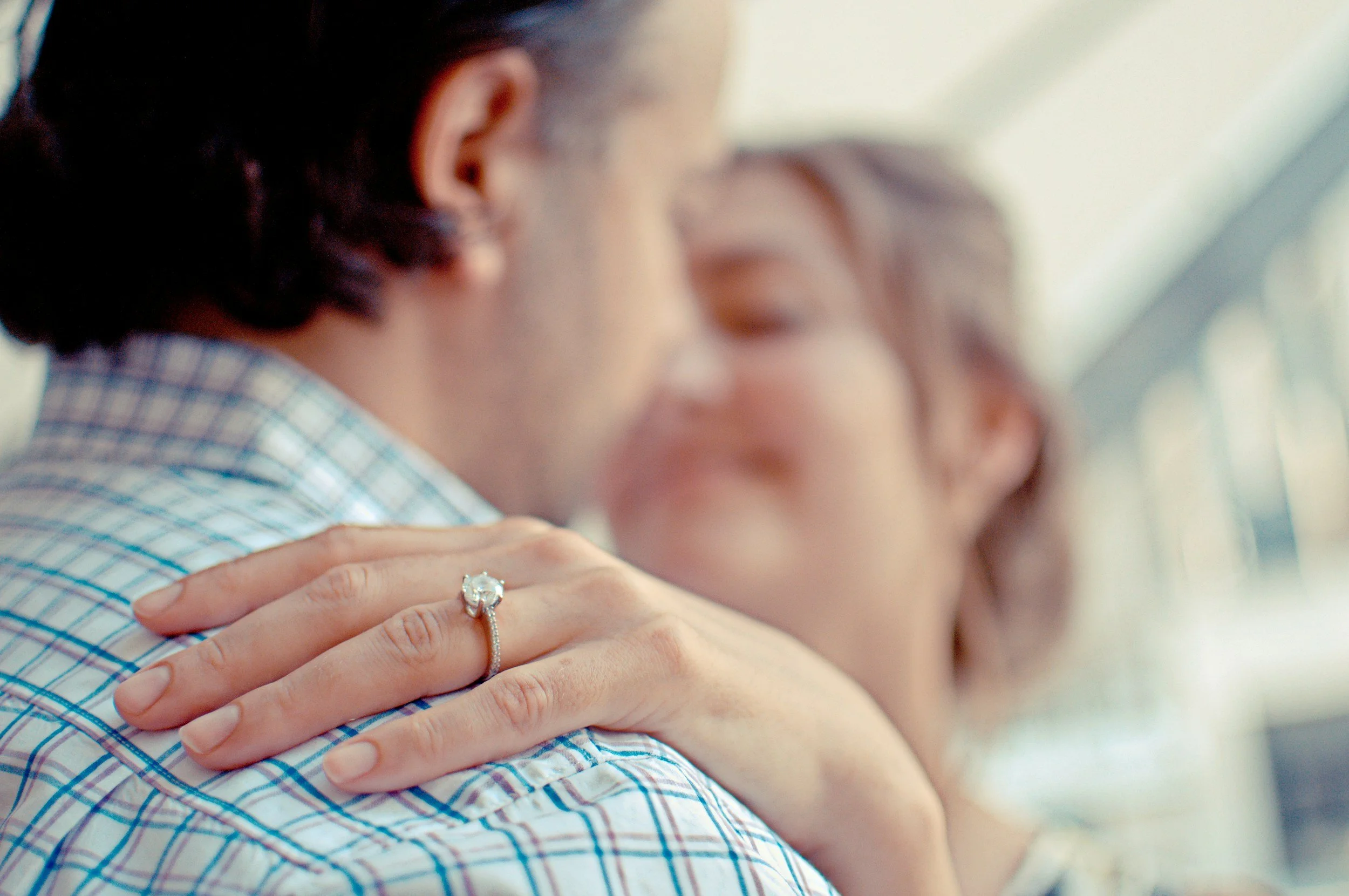 A close-up of a man and woman hugging, focusing on the woman's hand with a diamond ring on her finger resting on the man's shoulder. The woman has brown hair and the man has dark hair, both are slightly blurred in the background.