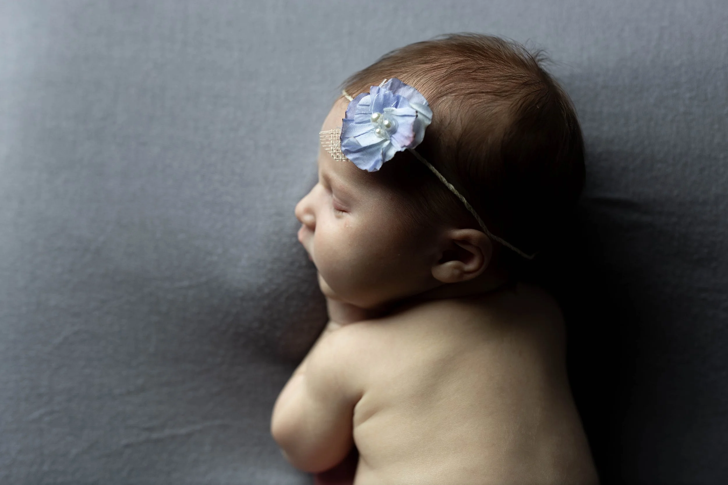 A sleeping newborn baby on a gray surface, wearing a headband with a light blue fabric flower and pearls.