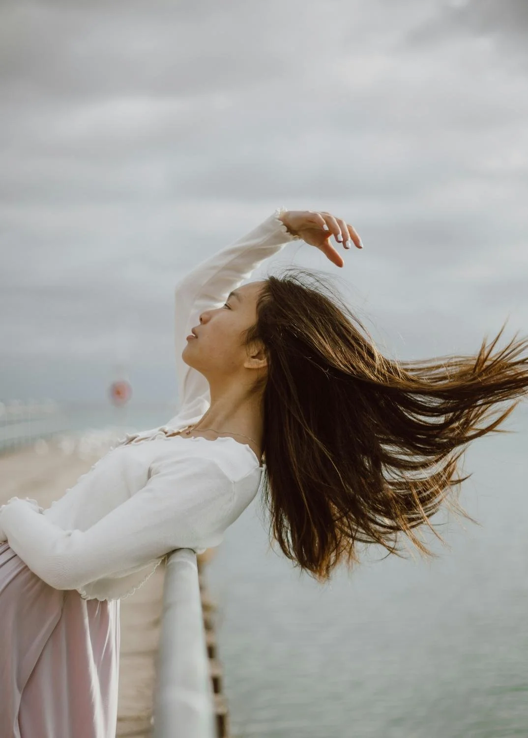 A woman with long brown hair standing on a pier near the water on a cloudy day.