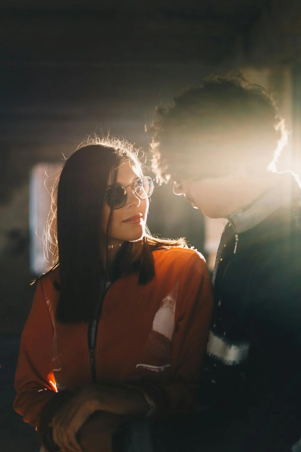 A woman wearing glasses and an orange jacket looking at a man with glasses, with sunlight behind them creating a backlit effect.