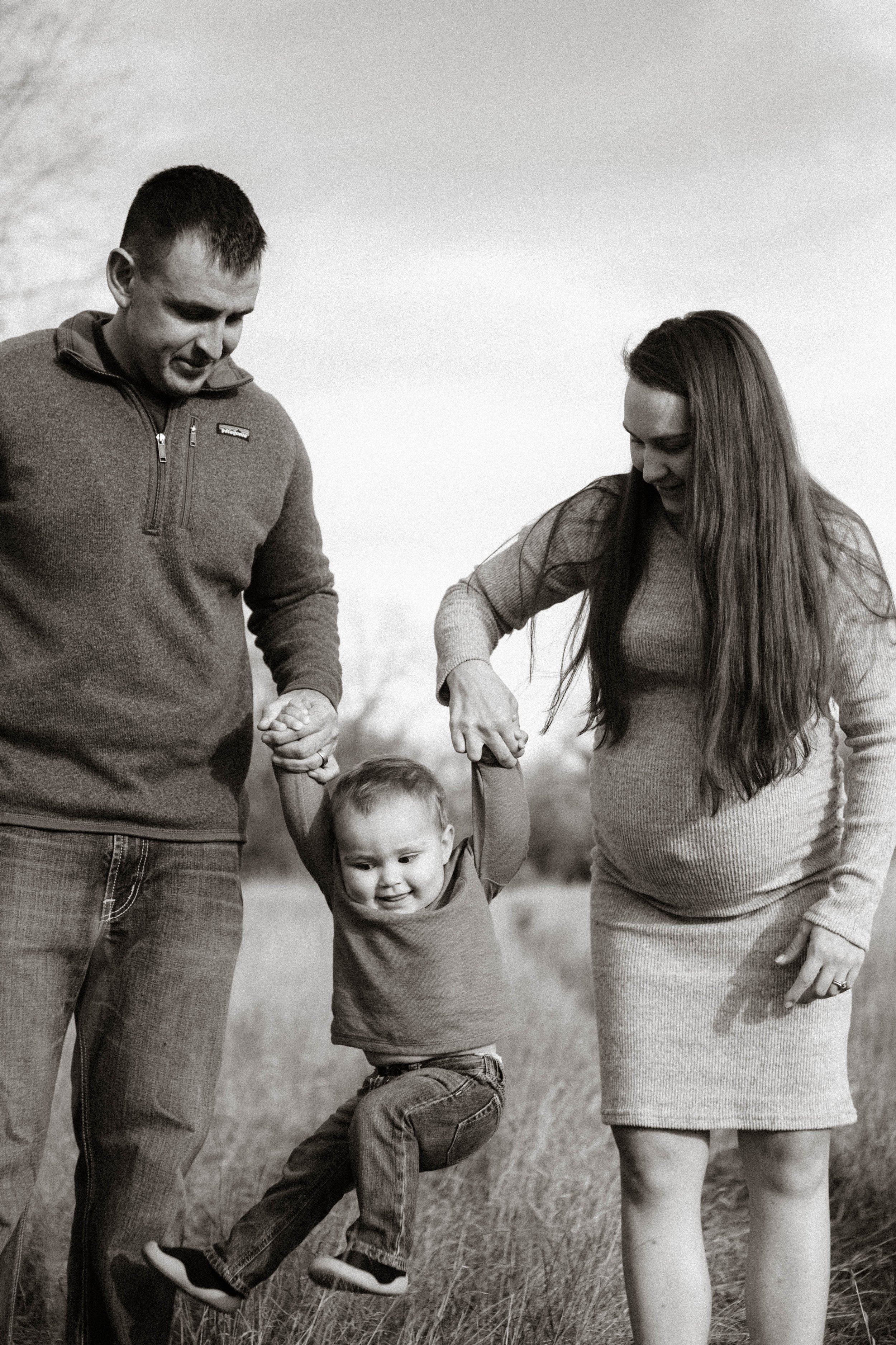 A happy family of three, including a toddler, walking outdoors in a grassy field, with the parents holding the child's hands and lifting him up as he smiles and swings his legs.