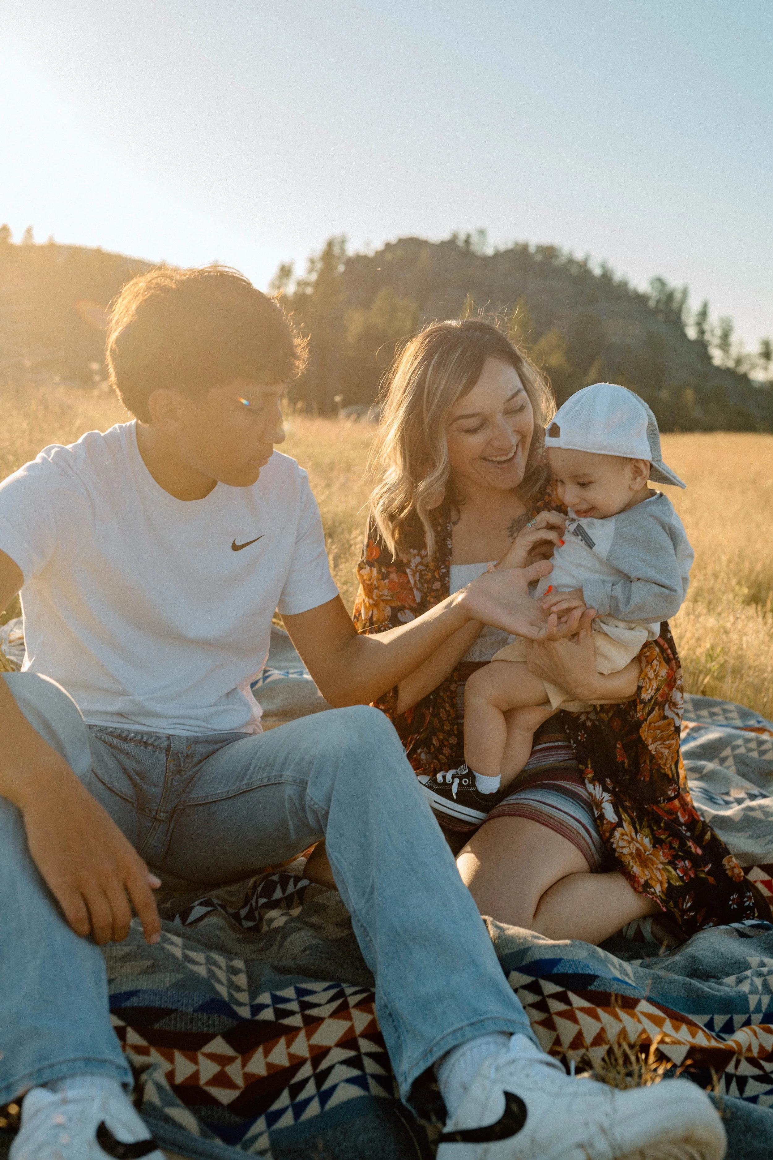 A woman and two children enjoying a picnic outdoors in a field with a hill in the background during sunset.