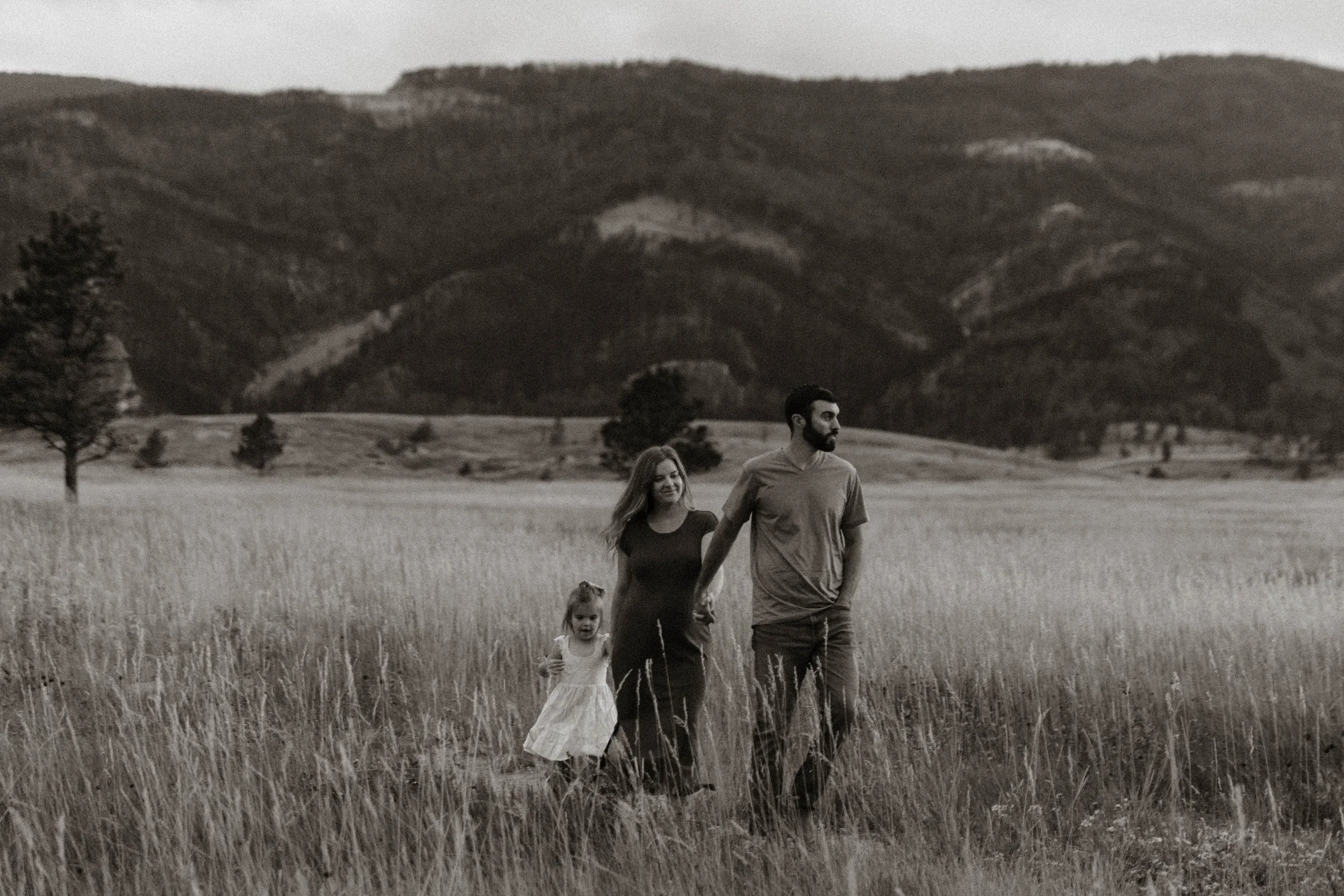 A family of three, including a man, a woman, and a young girl, walking through a grassy field with mountains in the background, in black and white.