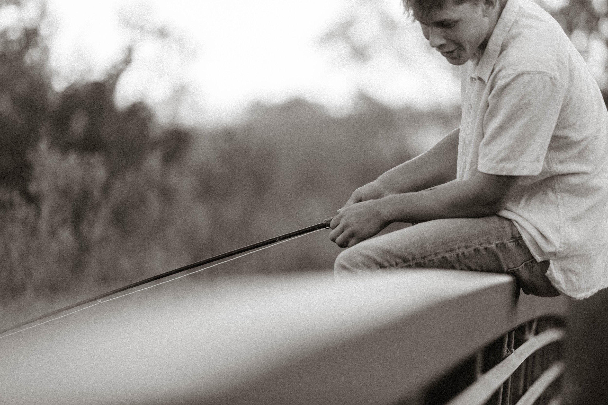 A person sitting on a bridge, fishing with a rod in an outdoor setting, captured in black and white.