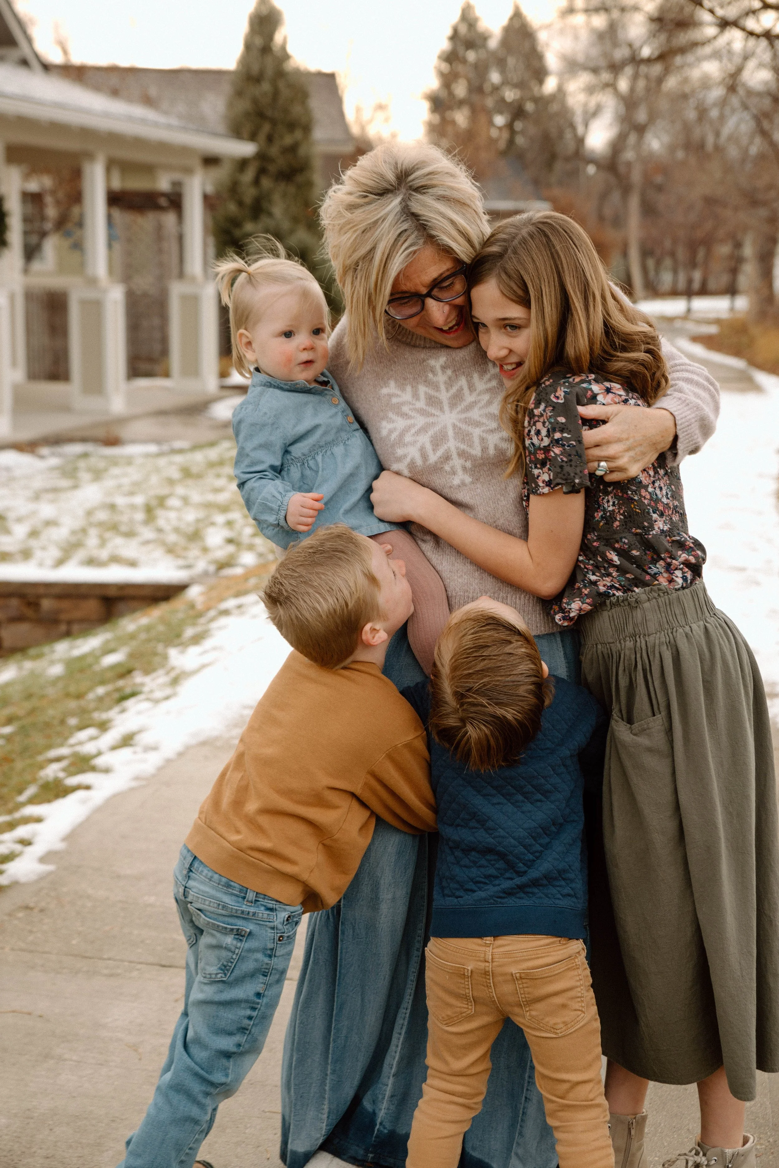 A woman and four children hugging outdoors on a chilly day with snow on the ground, smiling and showing affection.