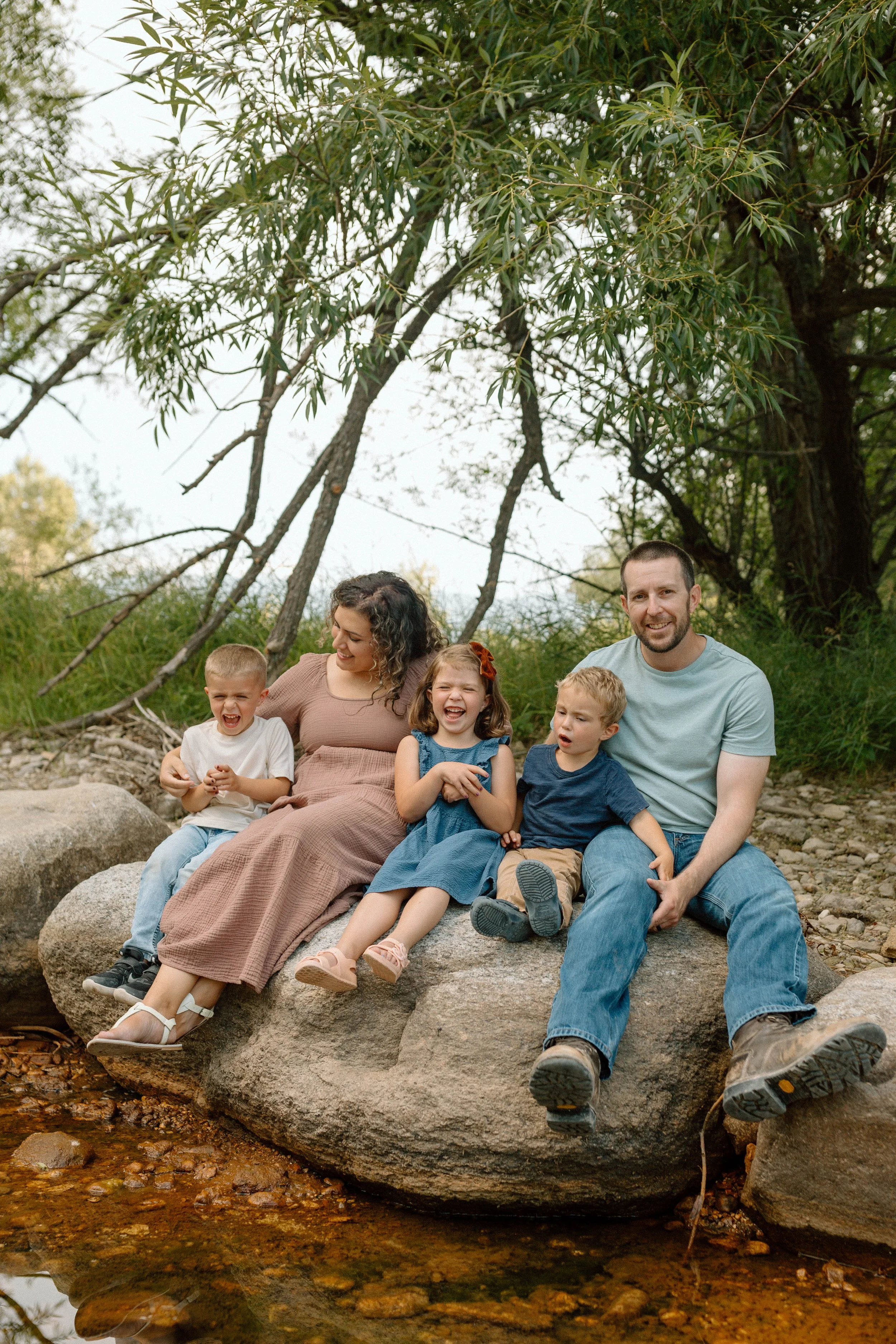 A family of five sitting on a large rock in a natural outdoor setting, smiling and laughing.