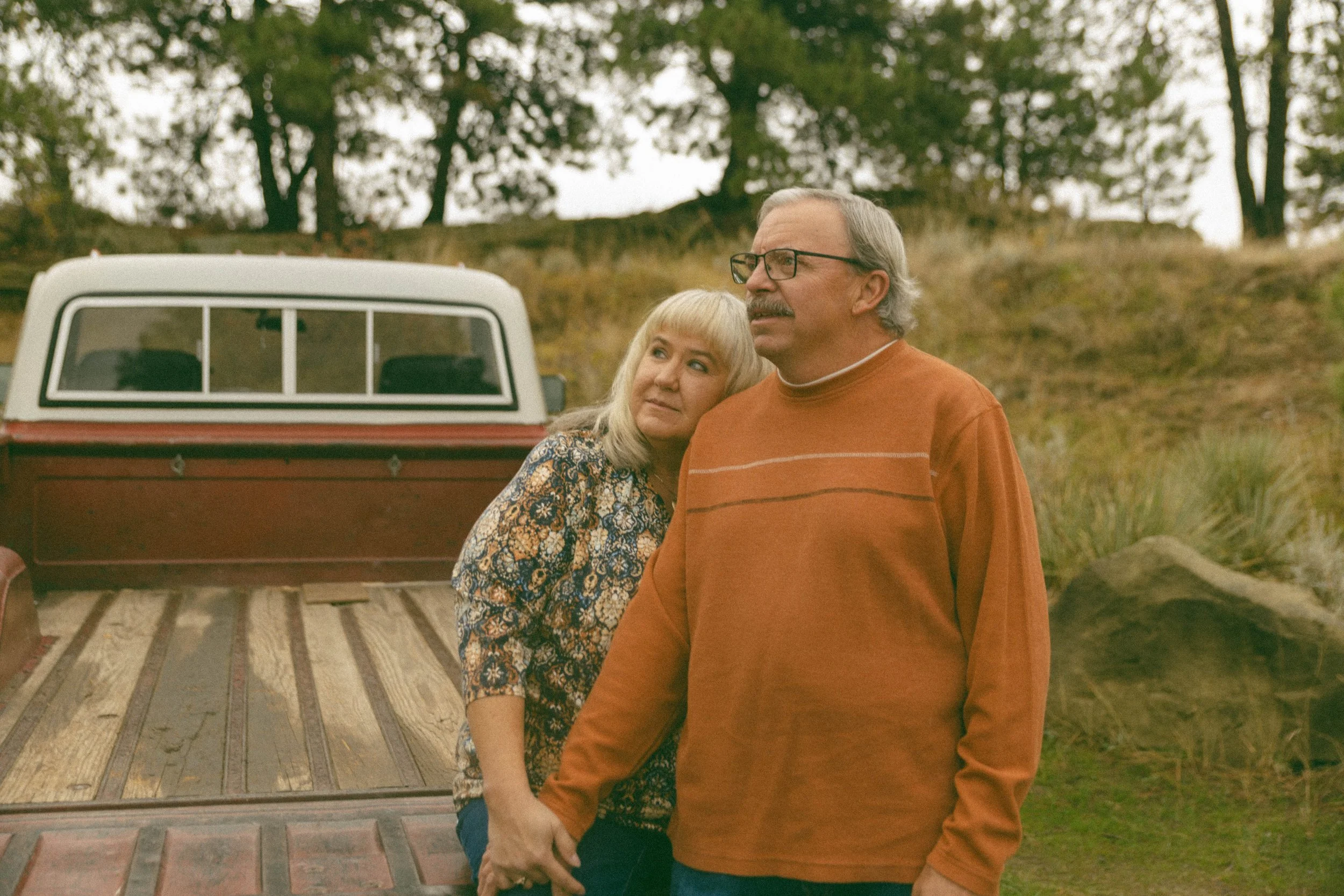 A middle-aged man with glasses and a mustache holding hands with a woman with blonde hair, standing outdoors near a red pickup truck, in a rural setting with trees and grass.