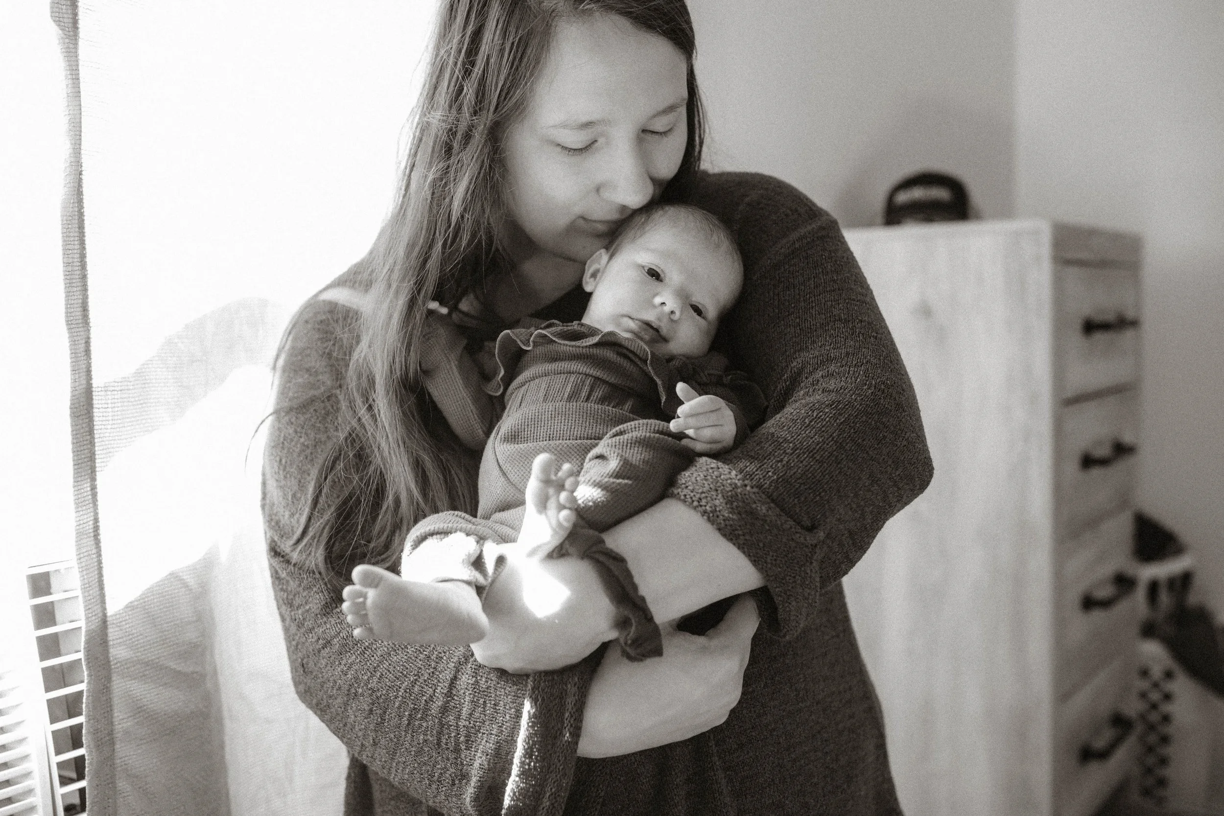 A woman holding a baby in her arms, standing near a window in a room.