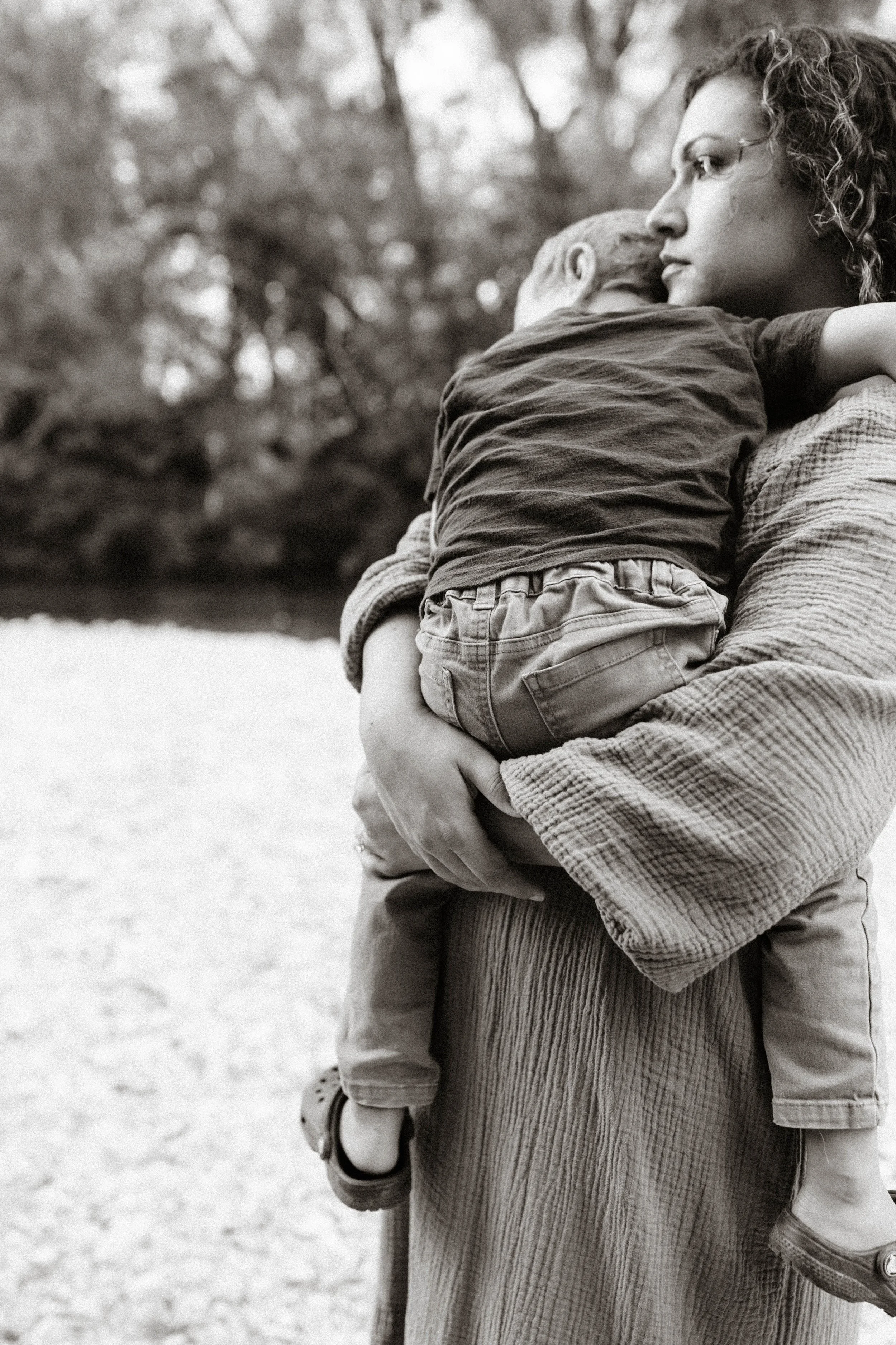 A woman holding a young boy outdoors near a body of water, with trees in the background, in black and white.