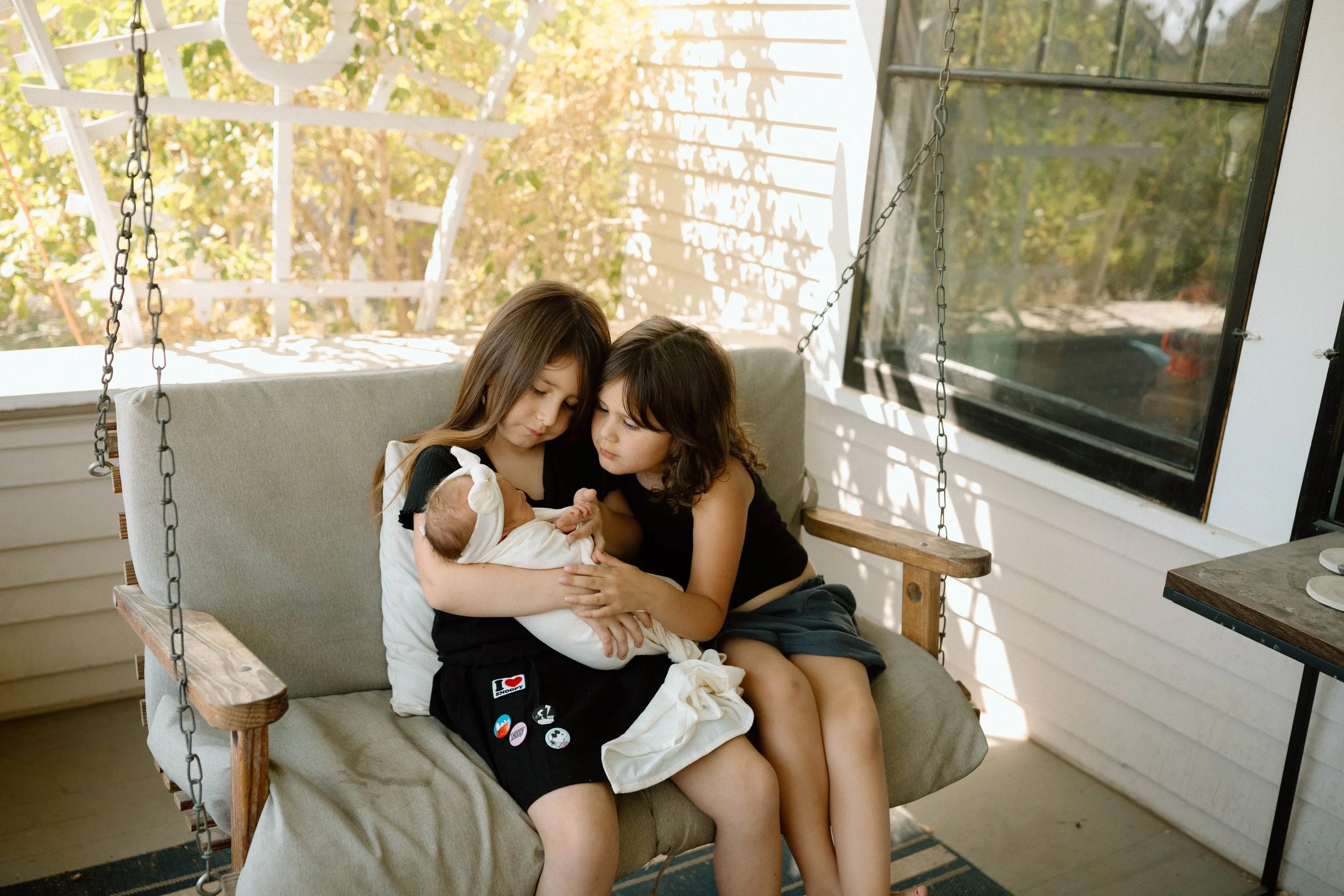 Two young girls sitting on a porch swing, holding and admiring a newborn baby girl wrapped in a white blanket, with a screened porch and outdoor trees in the background.