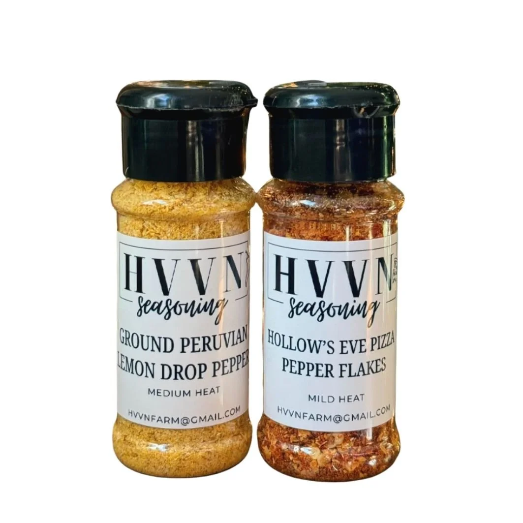 Two bottles of HVVN seasoning with black caps, one containing ground Peruvian lemon drop pepper and the other hollow's Eve pizza pepper flakes, against a white background.