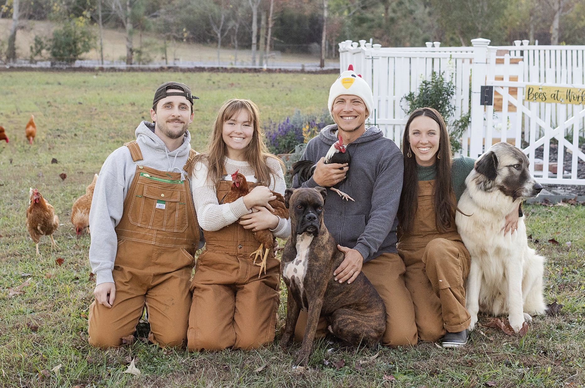 Group of four people and dogs on a farm with chickens, in front of a white fence with a yellow sign that reads 'Bees at Work'.
