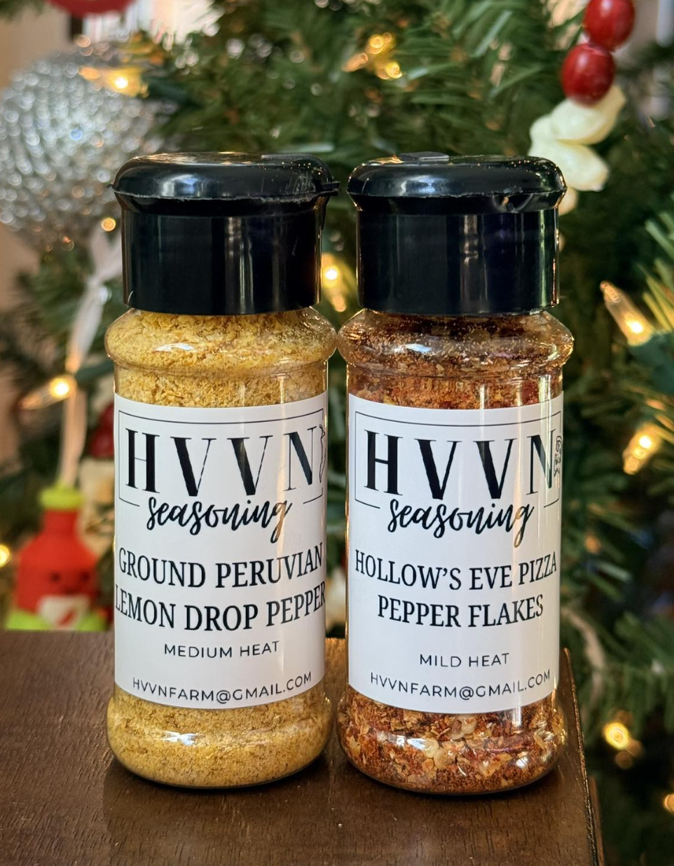 Two jars of HVVN seasoning in front of a decorated Christmas tree. The left jar contains ground Peruvian lemon drop pepper with medium heat, and the right jar contains Hollow's Eve pizza pepper flakes with mild heat.