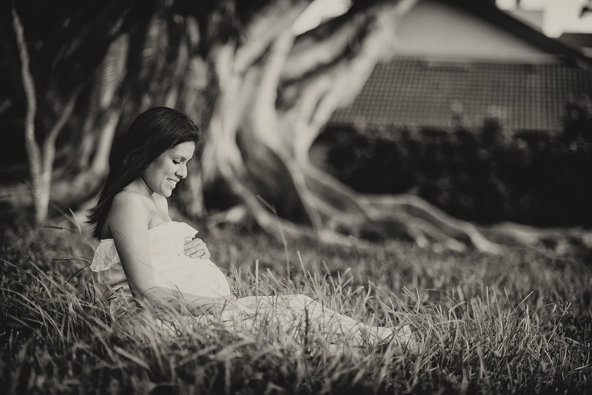 A pregnant woman sitting on the grass near a large tree, smiling and looking down, in black and white.
