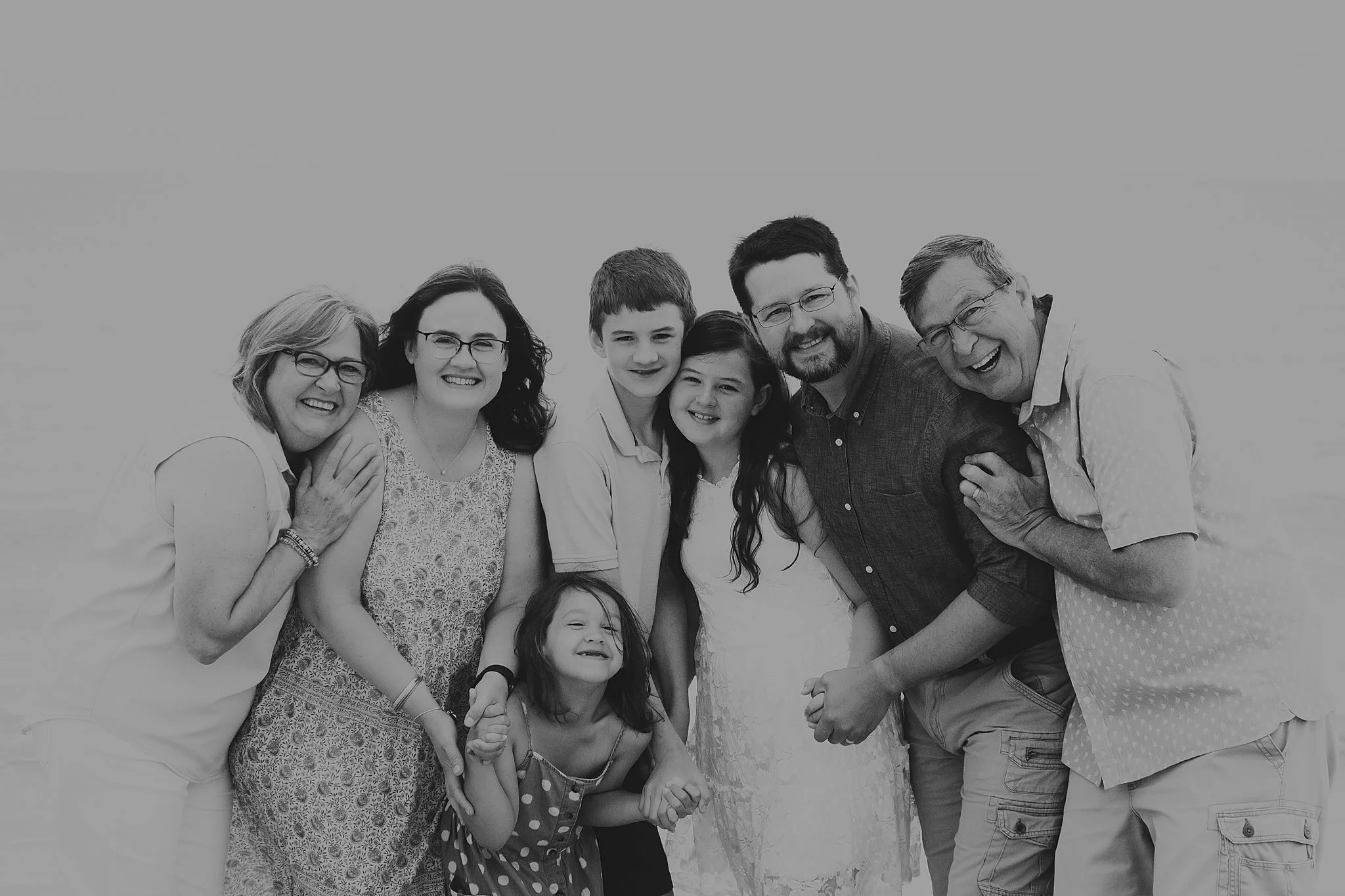 Black and white photo of a multi-generational family smiling together outdoors, including three women, two men, two boys, and one young girl.