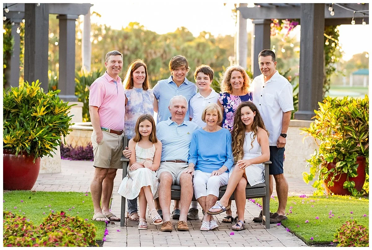 A multi-generational family photo outside during sunset, with 11 people, including grandparents, parents, and children, sitting on and standing around a bench in a garden with green plants and large potted bushes.