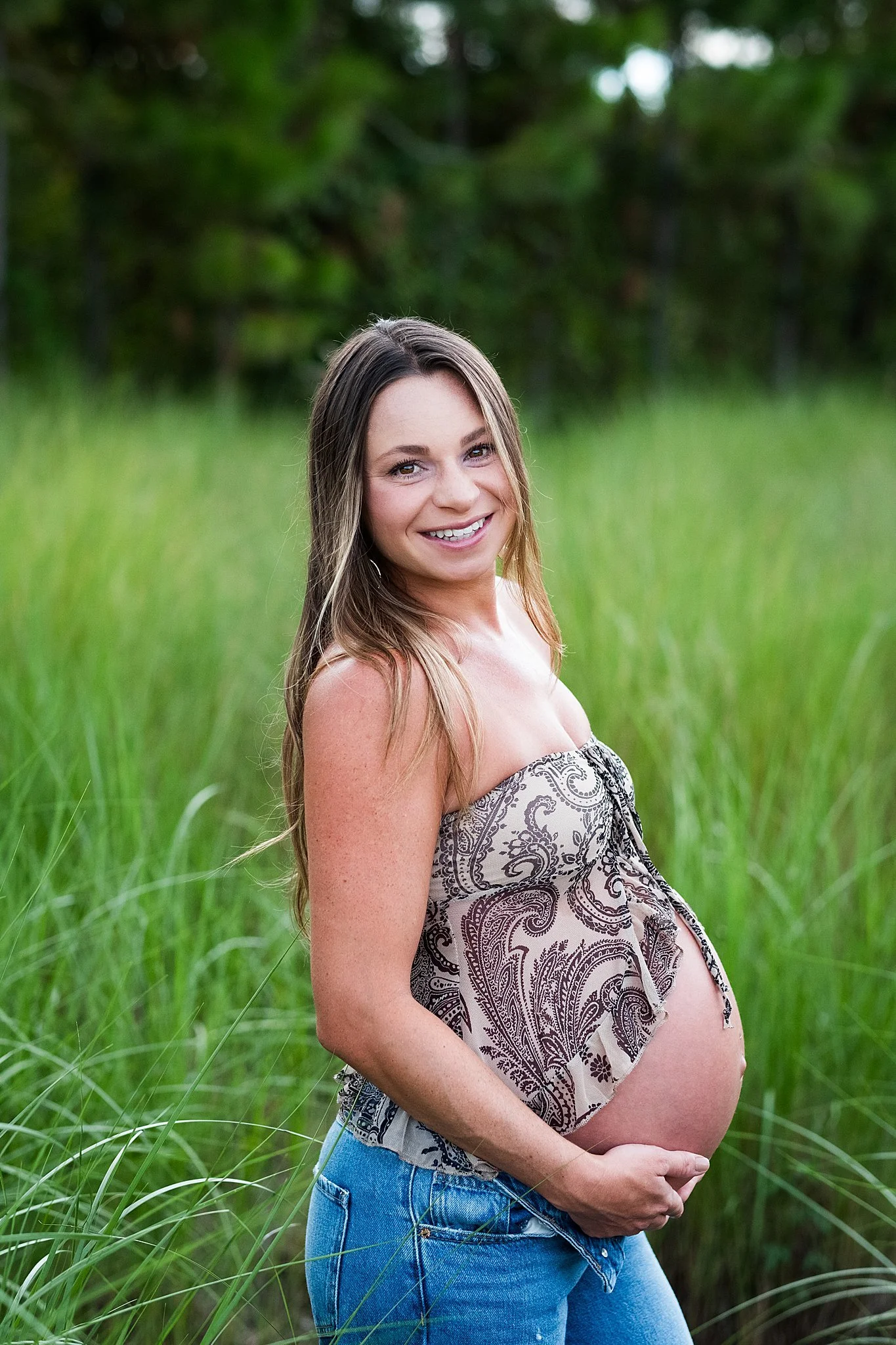 A pregnant woman smiling outdoors in a field of green grass, with trees in the background.