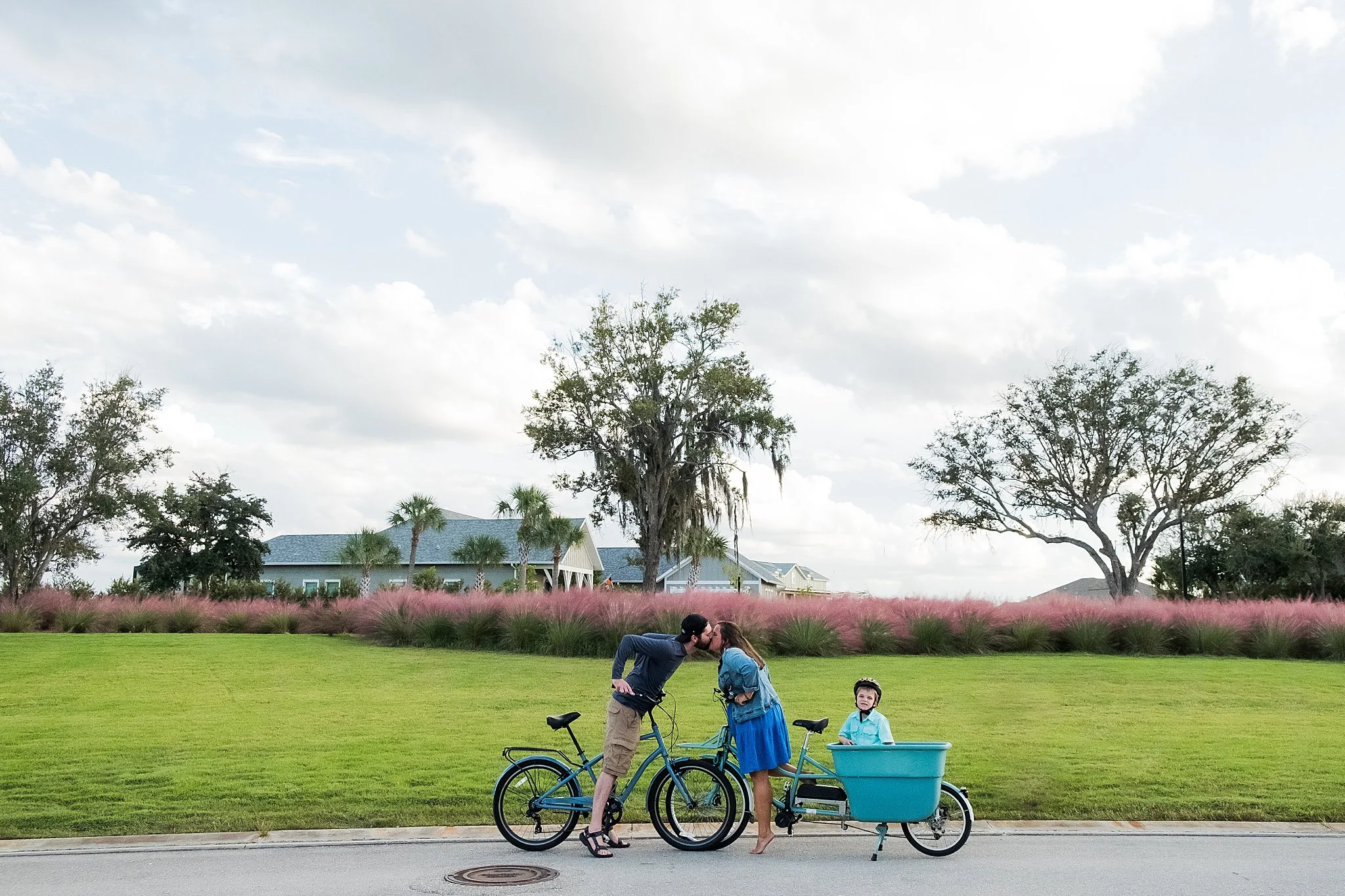 A couple sharing a kiss with a young child in a bike trailer on a suburban street, with trees and houses in the background.