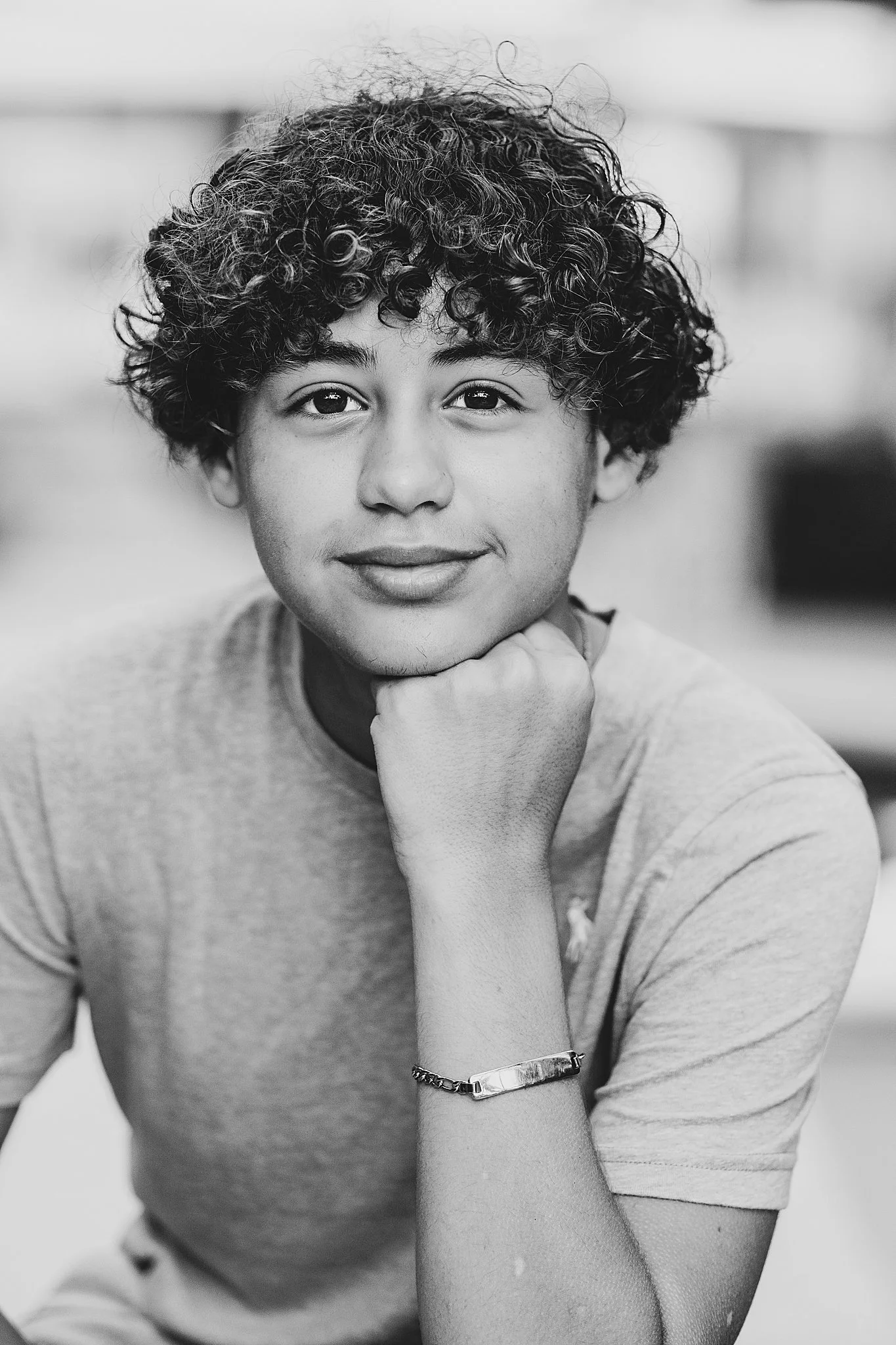 Black and white portrait of a young person with curly hair, smiling, propping chin on hand, wearing a short-sleeved t-shirt and bracelet.
